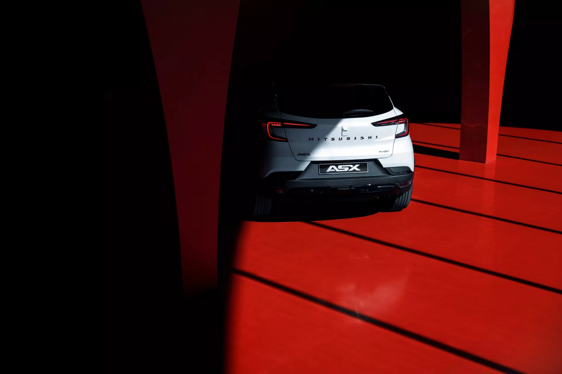 Zdjęcie z niskiego kąta tylnej części kompaktowego SUV-a Mitsubishi ASX 2023 z rzeźbionymi tylnymi światłami, chromowanym znaczkiem i tylną szybą, wykonane w dziennym świetle o wysokim kontraście na jaskrawoczerwonym podłożu.