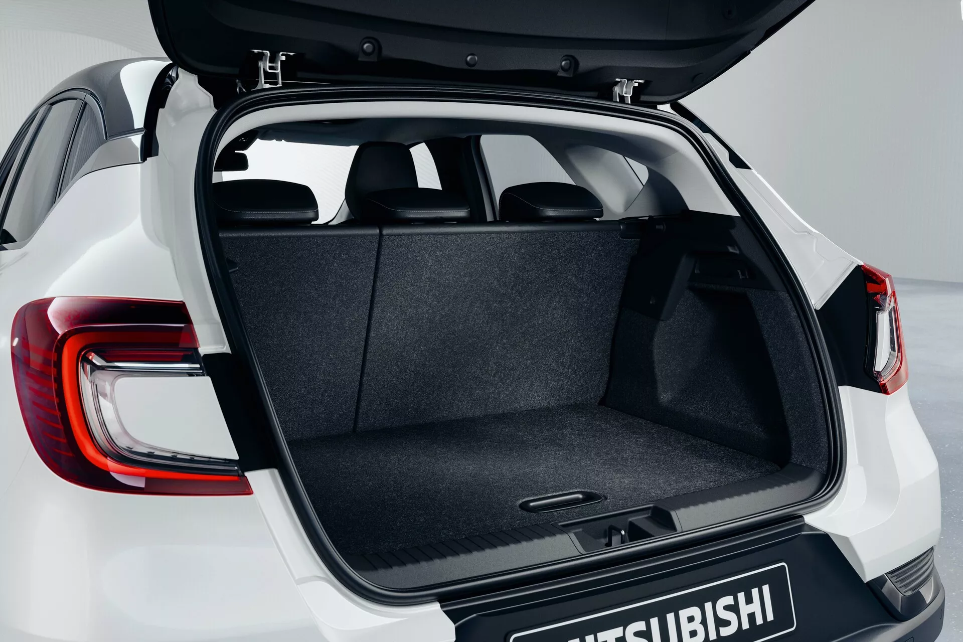 Przedział bagażowy Mitsubishi ASX 2023 (2. generacja) z otwartą klapą bagażnika widzianą pod lekko podwyższonym kątem, podkreślający wykładzinę bagażnika, mechanizm zawiasu, płaską podłogę oraz częściowo widoczne nowoczesne tylne światła LED z logo Mitsubishi na drzwiach bagażnika.