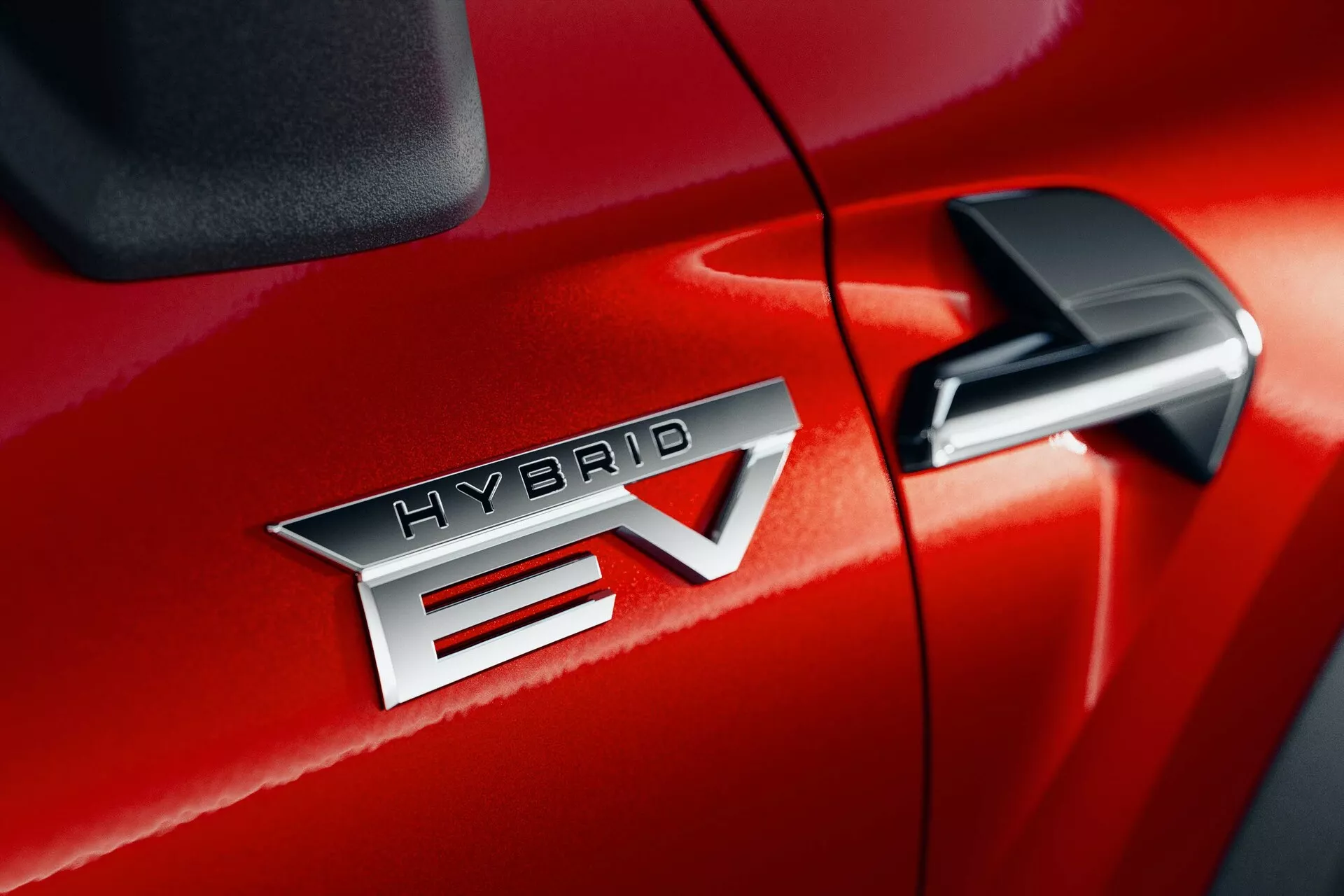 Zbliżenie detali Mitsubishi ASX 2023 (2. generacji) w czerwonym kolorze, ukazujące wyraźnie widoczny znaczek 'HYBRID EV', wskazujący na technologię hybrydowego pojazdu elektrycznego. Emblemat 'HYBRID EV' jest umieszczony centralnie, podkreślający metaliczne wykończenie i precyzyjne litery. 'EV' ma charakterystyczny strzałkowy wzór przecinający 'V'. Lusterko boczne jest częściowo widoczne w prawym górnym rogu, co sugeruje dodatkowe funkcje technologiczne. Lśniące wykończenie lakieru i wąska szczelina między przednimi drzwiami pasażera a przednim błotnikiem sugerują doskonałą jakość wykonania.