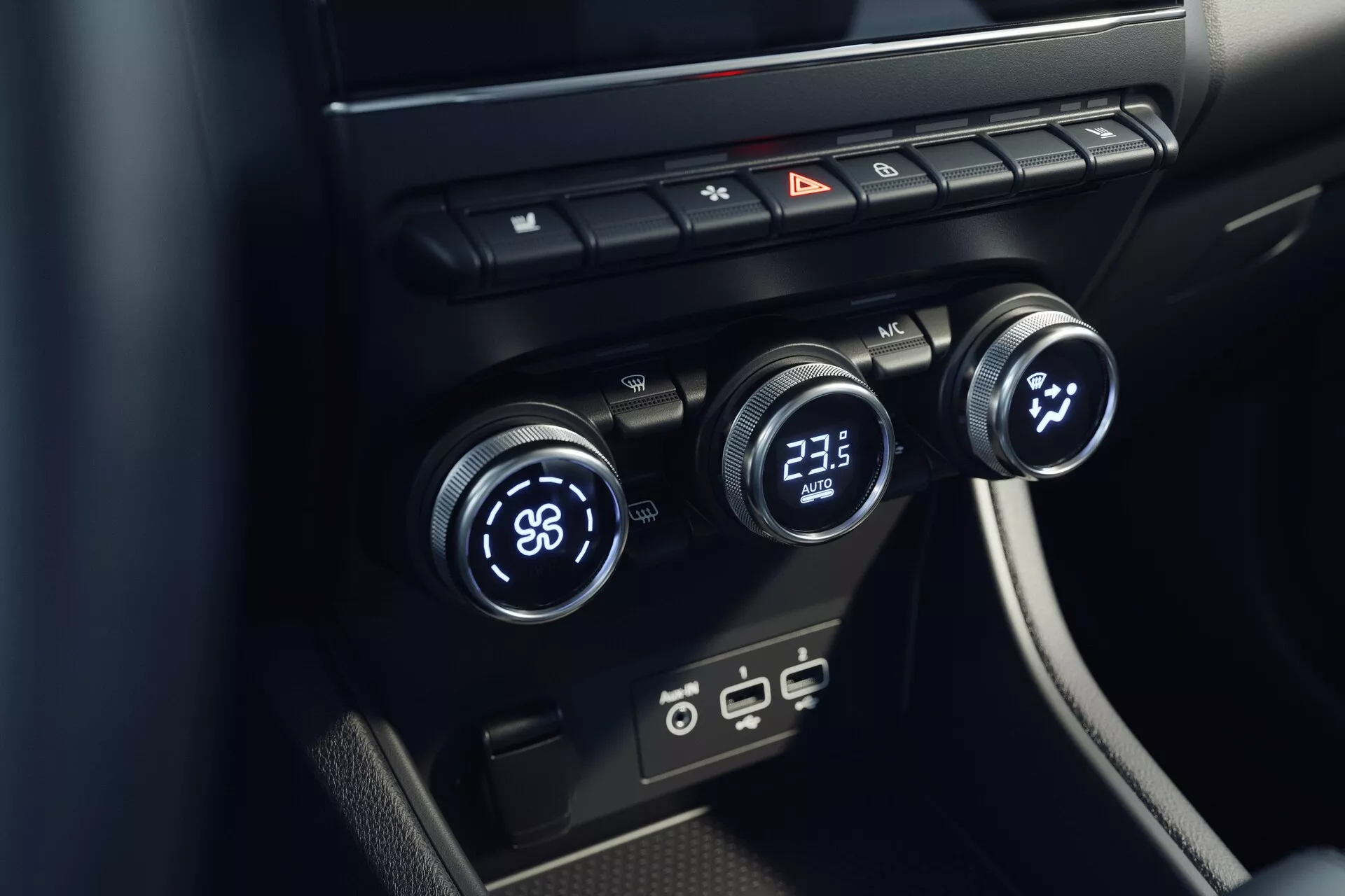 Zbliżenie systemu klimatyzacji wnętrza Mitsubishi ASX 2023 (2. generacja). Zdjęcie zrobione pod podwyższonym kątem skupia się na centralnej konsoli z trzema pokrętłami do regulacji temperatury: lewe ustawione na 'LO' a prawe na '23.5'. Środkowe pokrętło prawdopodobnie kontroluje prędkość lub kierunek wentylatora. Powyżej są przyciski do odmrażania, świateł awaryjnych i cyrkulacji powietrza. Poniżej znajdują się nowoczesne porty USB. Design jest minimalistyczny z eleganckimi czarnymi powierzchniami.