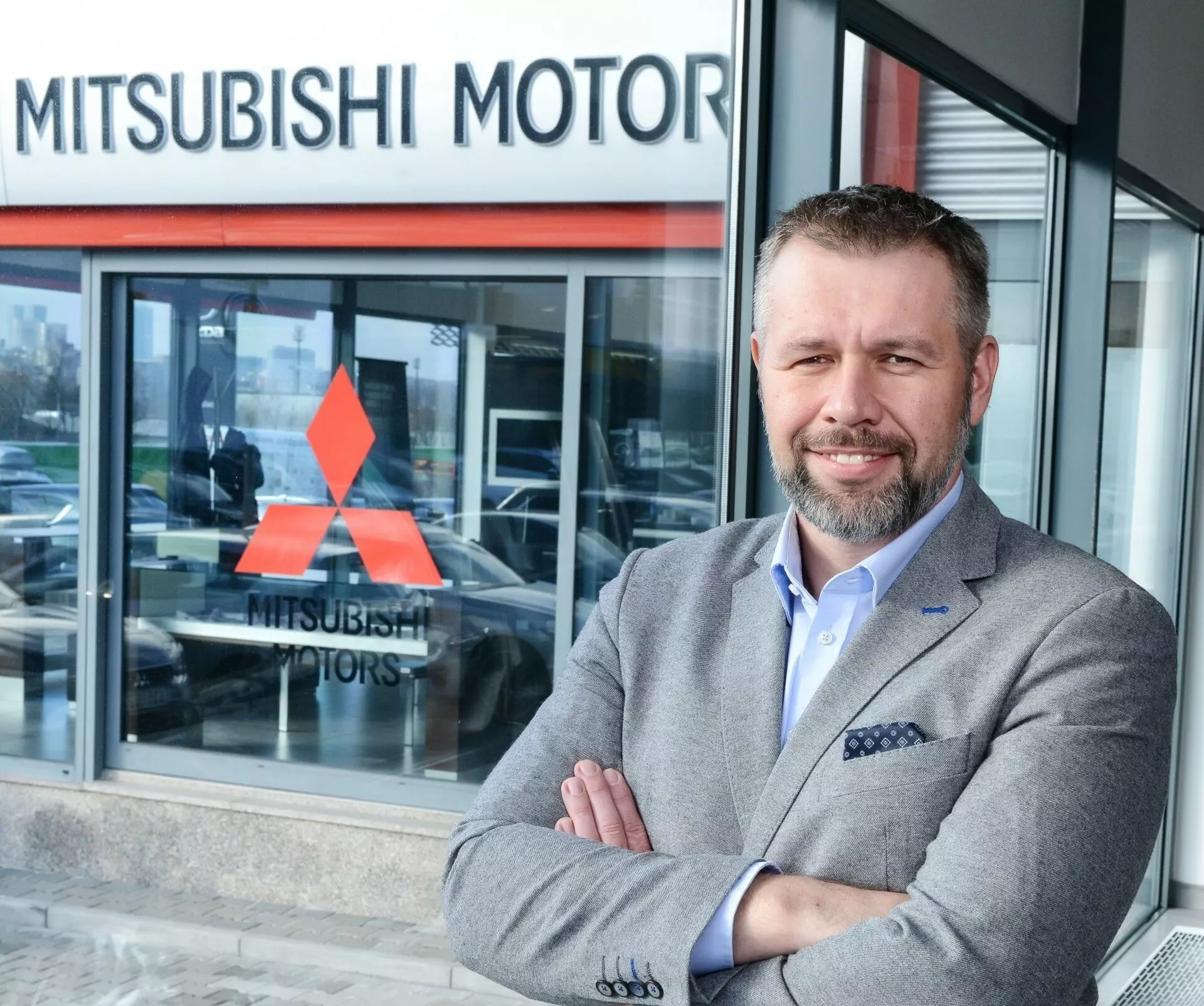 Osoba stojąca ze skrzyżowanymi rękami przed salonem Mitsubishi Motors, częściowo widoczny pojazd przez szybę sugerujący model wystawowy w tle