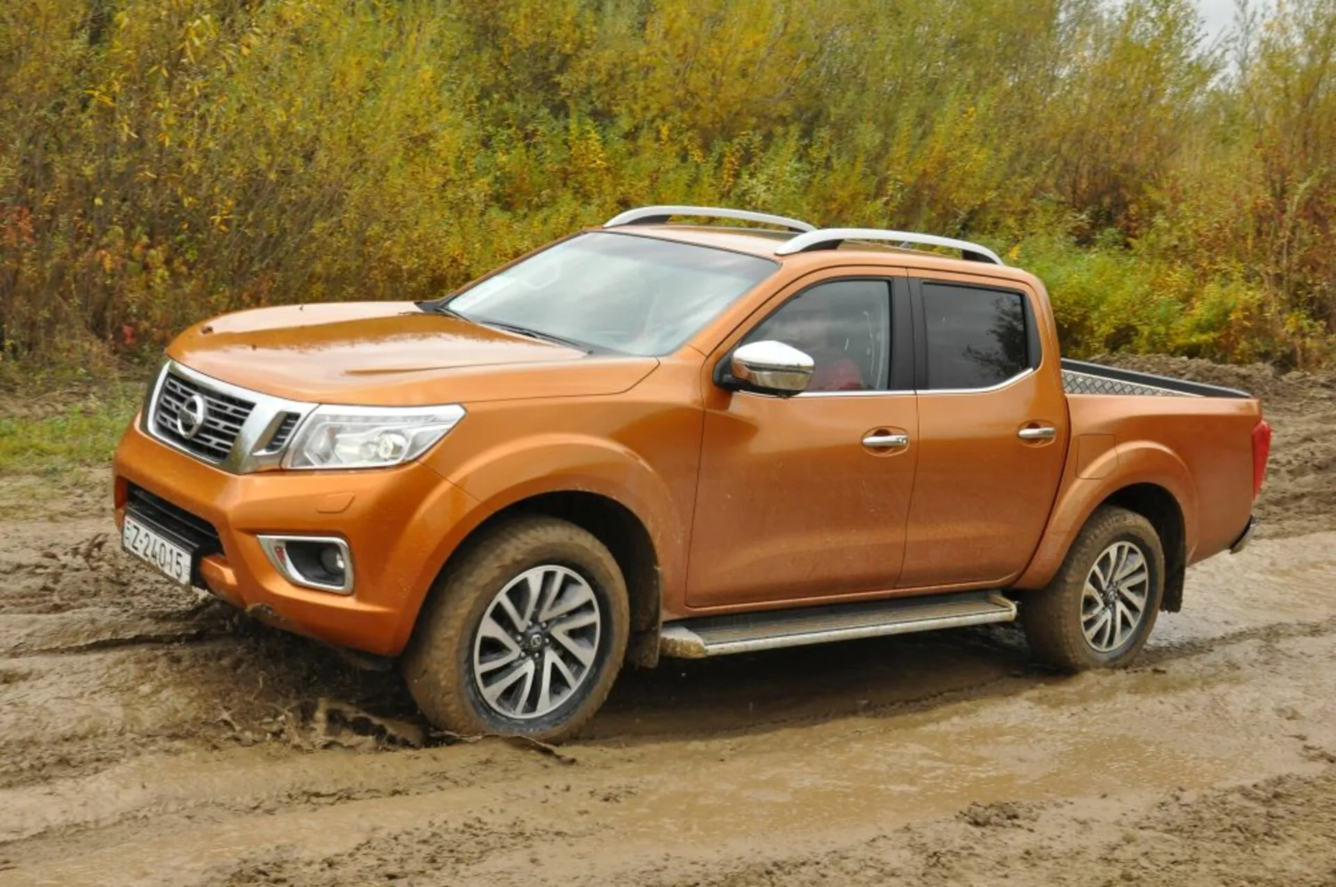 Pomarańczowy pickup Nissan Navara z 2016 roku w widoku trójkwartalnym z przodu, prezentujący reflektory, atrapę z akcentami chromowymi, koła ze stopów lekkich i konfigurację kabiny załogi na nierównym terenie