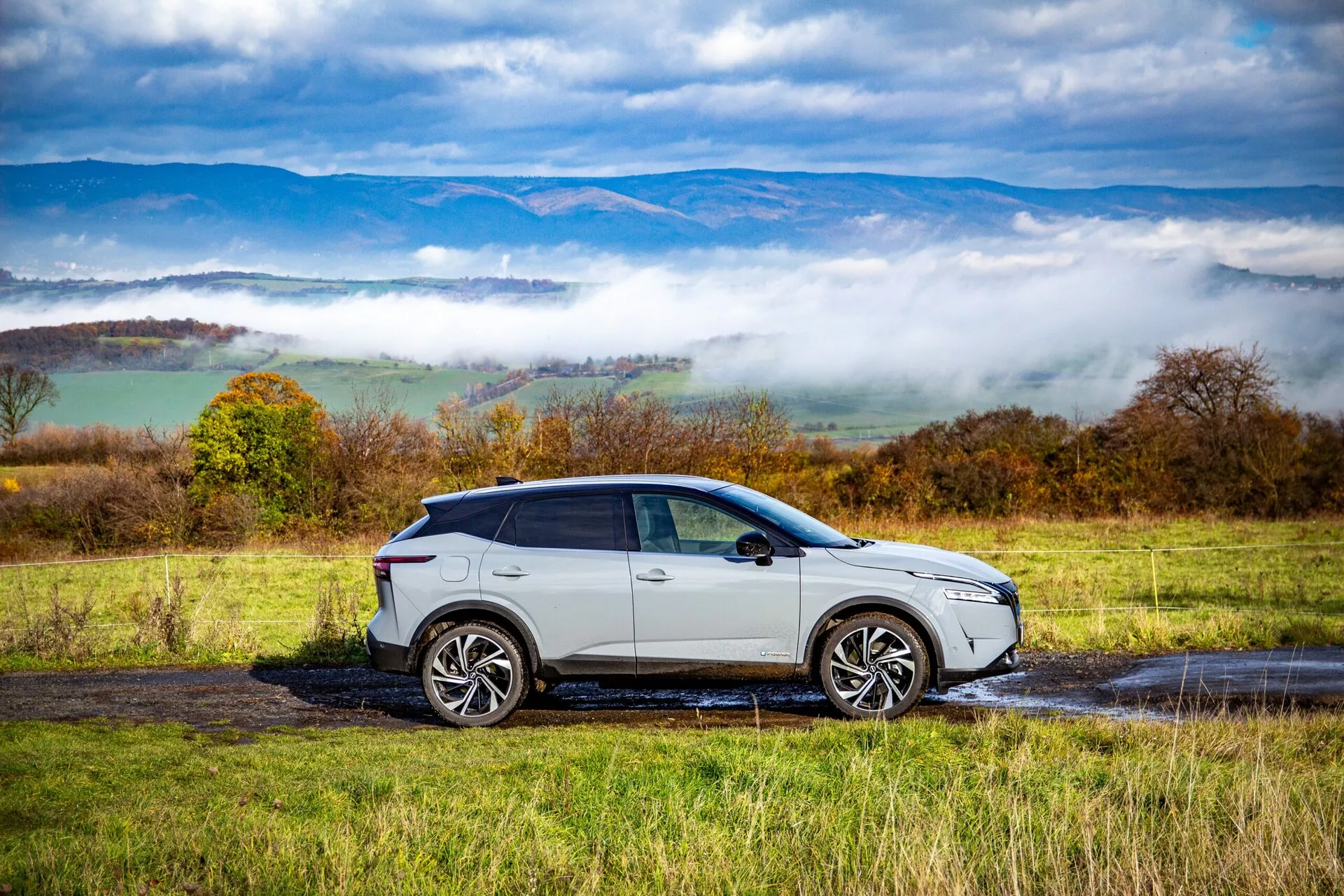Widok boczny profilu Nissana Qashqai 2021, nowoczesnego SUV-a z połyskującym srebrnym lub jasnoszarym wykończeniem, dużymi wieloramiennymi felgami aluminiowymi, przyciemnionymi szybami i opadającą linią dachu, zaparkowanego na mokrej wiejskiej drodze z kałużami. Elegancki design samochodu, nowoczesne jednostki oświetleniowe i zintegrowane kierunkowskazy w lusterkach bocznych są wyraźnie widoczne na tle falujących wzgórz i nisko zawieszonych chmur.