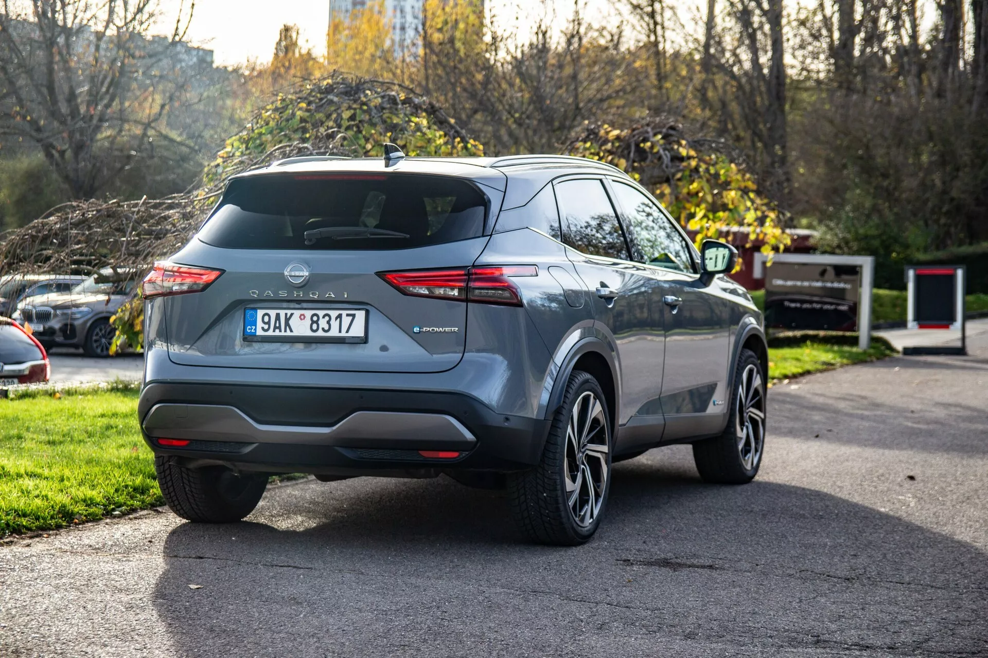 Samochód Nissan Qashqai 2021 sfotografowany pod kątem trzech czwartych z tyłu, ukazujący charakterystyczne tylnie światła, widoczny tylny bagażnik z wycieraczką, centralnie umieszczony znak producenta, zamontowaną tablicę rejestracyjną, zintegrowane odblaski w tylnym zderzaku, pojedynczą końcówkę wydechu, felgi aluminiowe, nowoczesne linie nadwozia, zwężające się boczne okna, potencjalne relingi dachowe, krótką antenę dachową, metaliczny lakier i częściowo widoczne boczne lusterko.