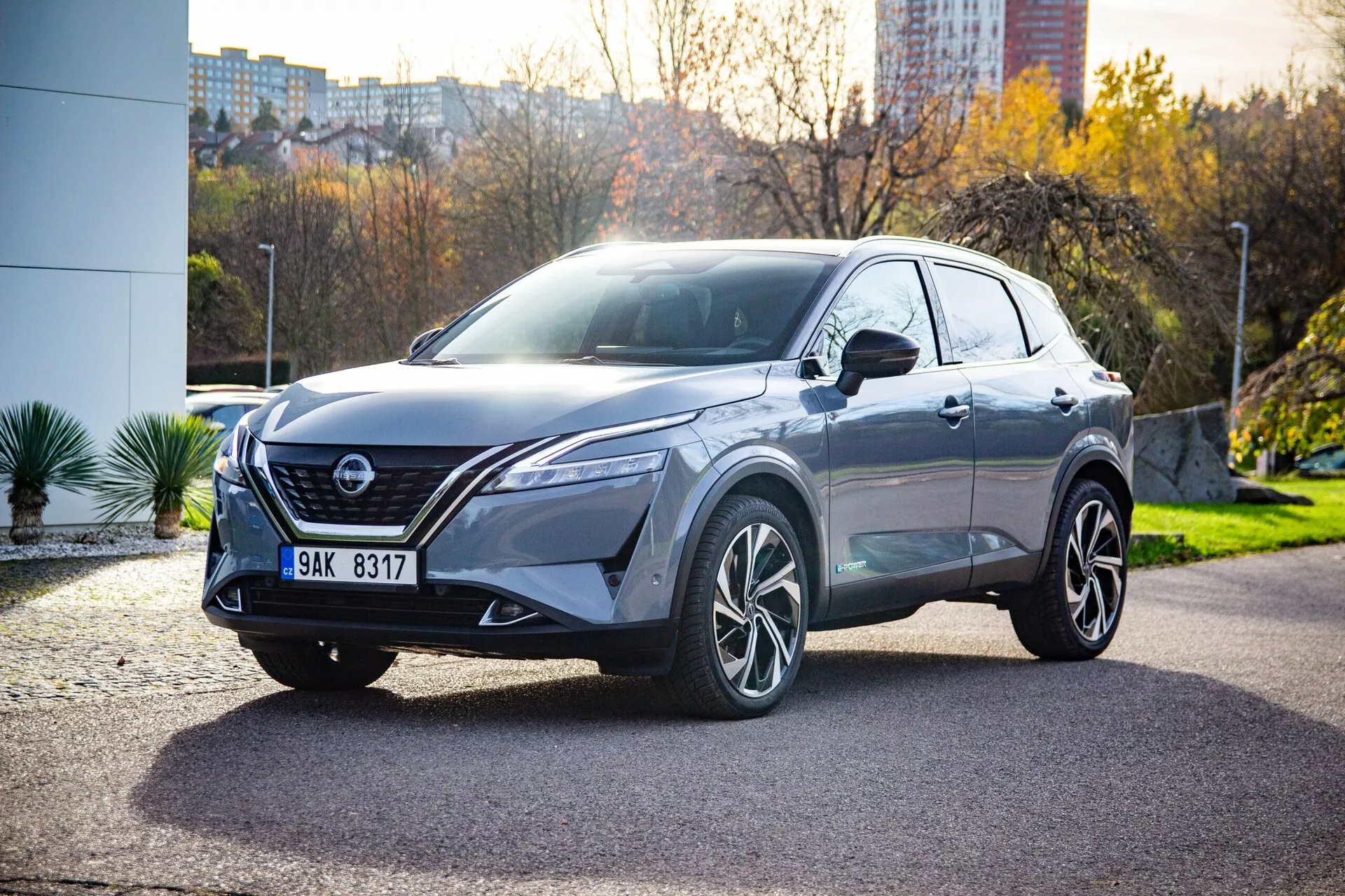 Trzy-czwarte przedniego widoku nowoczesnego crossovera SUV Nissan Qashqai 2021, ustawionego pod kątem na utwardzonym terenie z miejskim tłem zieleni i budynków. Widoczne cechy kluczowe obejmują eleganckie, kątowe reflektory LED, wyraźną kratkę w kształcie litery V, zintegrowane światła przeciwmgielne, czujniki parkowania, rzeźbioną maskę, wyraźne łuki kół, wieloszprychowe felgi ze stopu, sensoryczne komponenty na kratce dla zaawansowanych systemów wspomagania kierowcy, kolorowe lusterka boczne najprawdopodobniej z zintegrowanymi kierunkowskazami, panoramiczny dach, przyciemniane szyby, sugerujące siedzenia pokryte skórą oraz aerodynamiczne linie nadwozia.