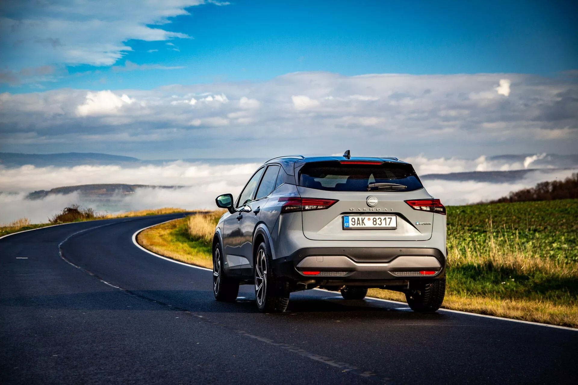 SUV Nissan Qashqai 2021 widziany pod tylnym kątem trzech czwartych na wijącej się wiejskiej drodze pod rozproszonymi chmurami, prezentujący elegancki i nowoczesny design z wąskimi poziomymi tylnymi światłami, dyskretnymi rurami wydechowymi, wieloramiennymi felgami z lekkich stopów, relingami dachowymi i zaawansowanymi czujnikami parkowania. Jasny metaliczny kolor podkreśla jego luksusowy charakter.