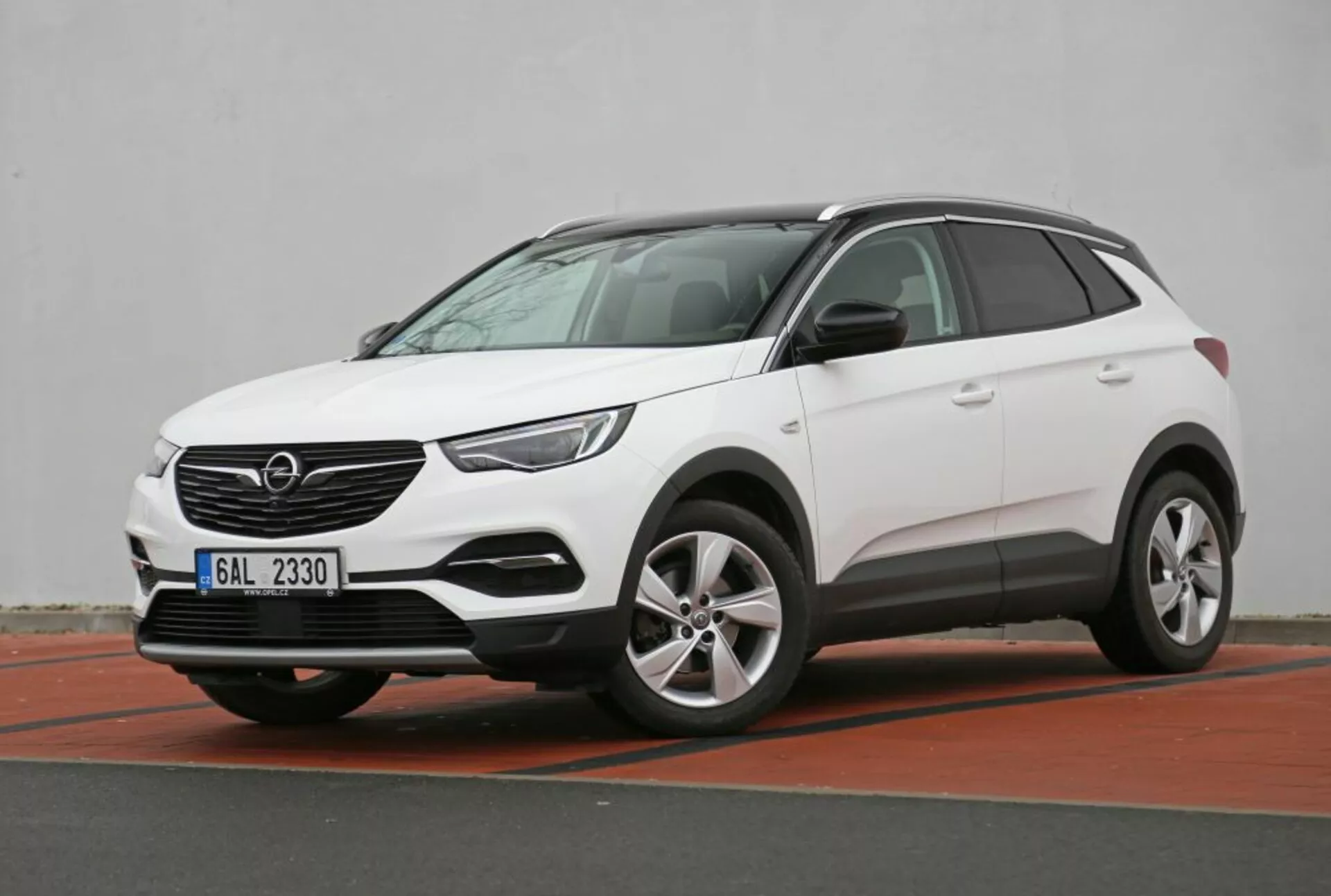 Białe kompaktowe SUV crossover Nissan Qashqai z 2014 roku zaparkowane pod kątem prezentujące swoje profile przednie i boczne, podkreślając jego wysokość jazdy z niskiego kąta, nowoczesne reflektory LED, czujniki parkowania na przednim zderzaku z iluzją płyty osłonowej i felgi aluminiowe, w miejskim otoczeniu parkingowym z czerwonym krawężnikiem