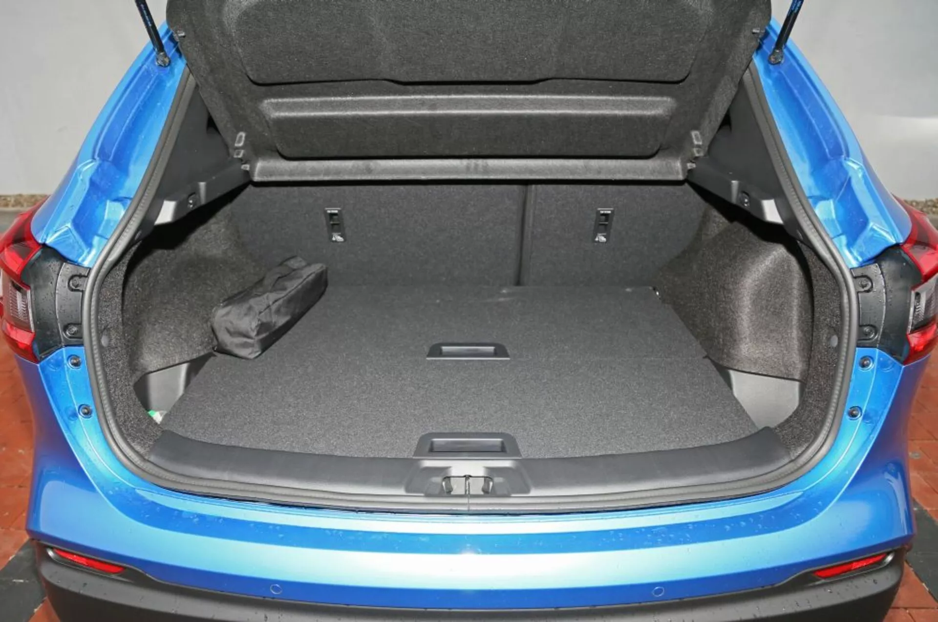 Widok na tylny bagażnik hatchbacku Nissan Qashqai 2014 z otwartymi drzwiami, pokazujący czystą szarą wykładzinę, czarny rulon lub torbę położoną z boku, żywe niebieskie panele wewnętrzne i funkcje do rozbudowy przestrzeni bagażowej