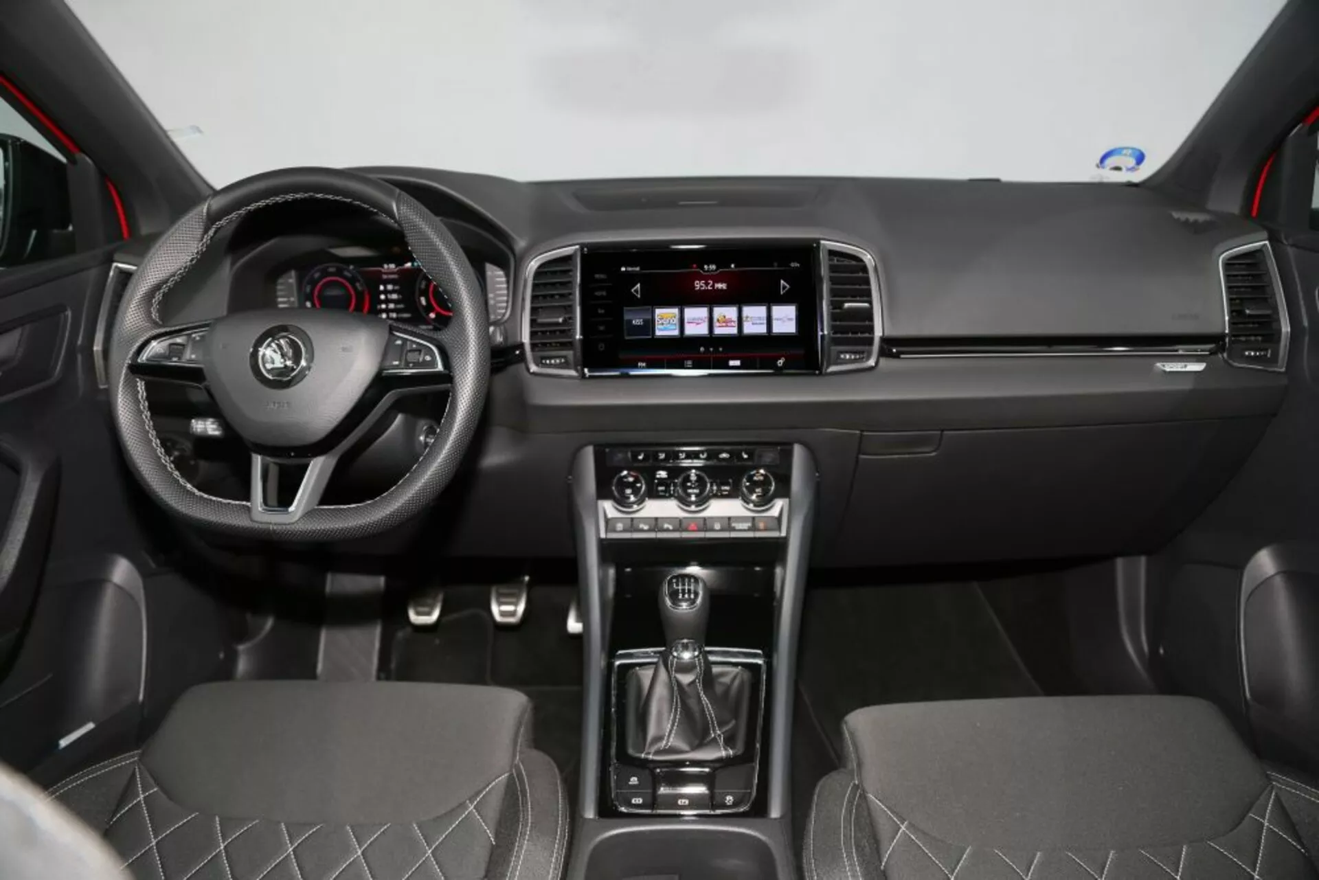 Widok frontalny wnętrza Nissan Qashqai 2014 pokazujący deskę rozdzielczą, kierownicę z przyciskami sterującymi, system infotainment, manualną dźwignię zmiany biegów, kontrolę klimatyzacji i siedzenia pokryte tkaniną z diamentowym pikowaniem.