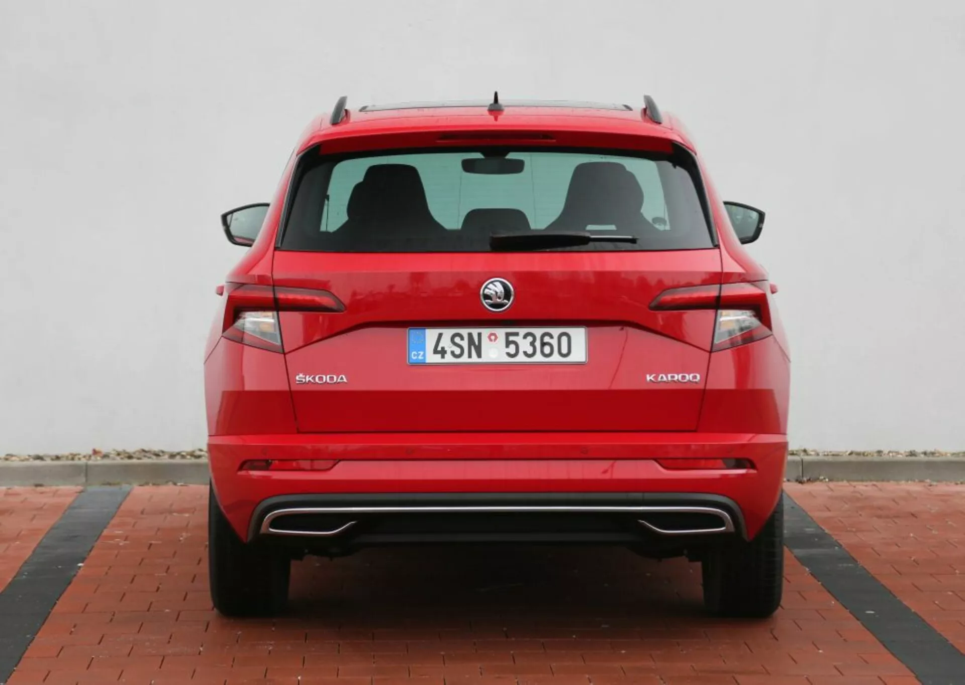 Szczegółowy widok tylnej części czerwonego Skoda Kodiaq 2014, pokazujący symetryczny tylny design z logiem Skoda, kryształowe tylne światła, tablicę rejestracyjną, czujniki parkowania na zderzaku, podwójne końcówki wydechu i płetwę rekina antenę, na neutralnym tle z wzorzystym brukiem.