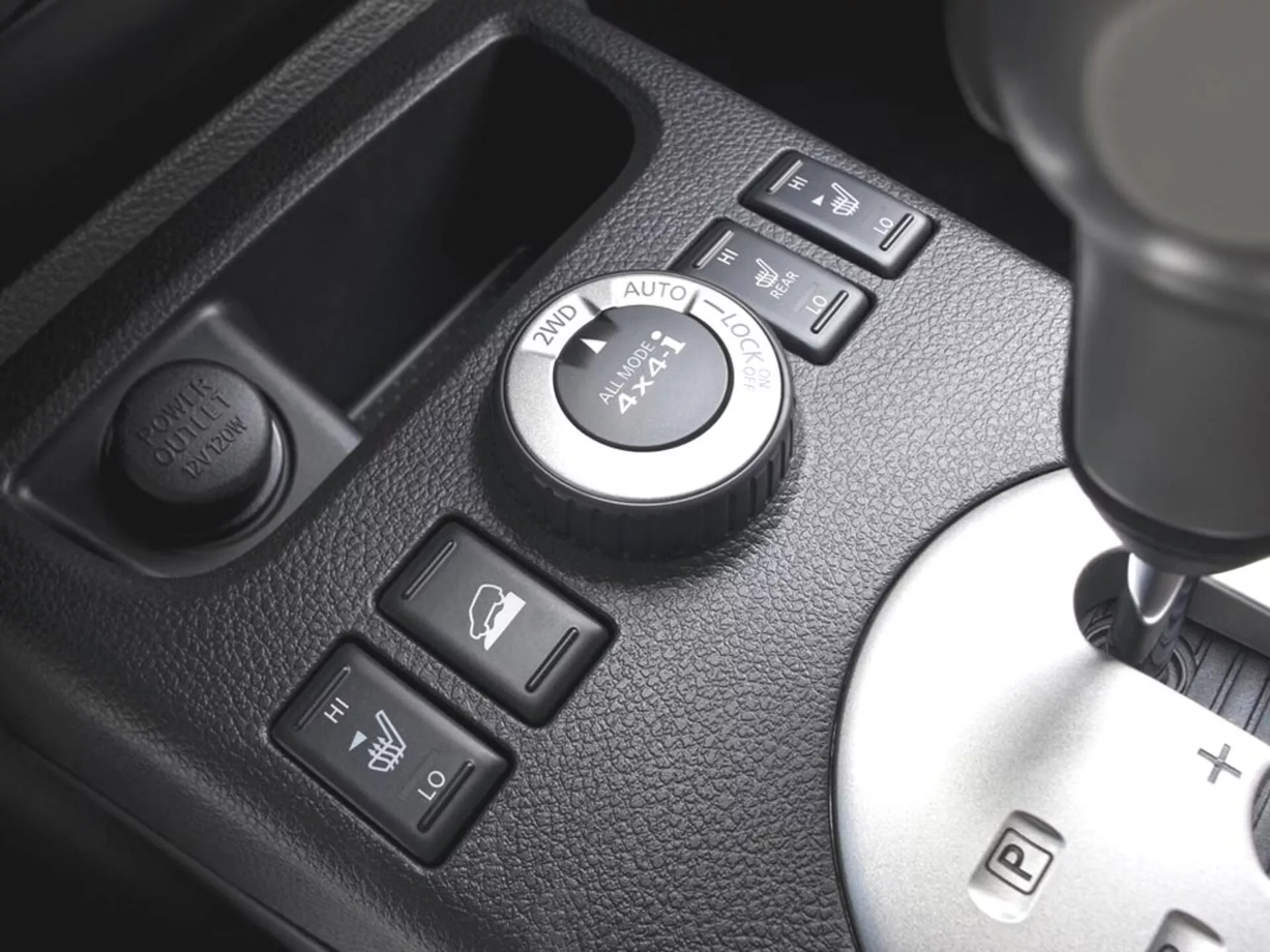 Zbliżony widok na konsolę środkową Nissan X-Trail 2010 z pokrętłem kontrolnym 4WD, przyciskami podgrzewania siedzeń, przyciskiem gniazdka zasilającego i dźwignią zmiany biegów z opcją manualnego przełączania