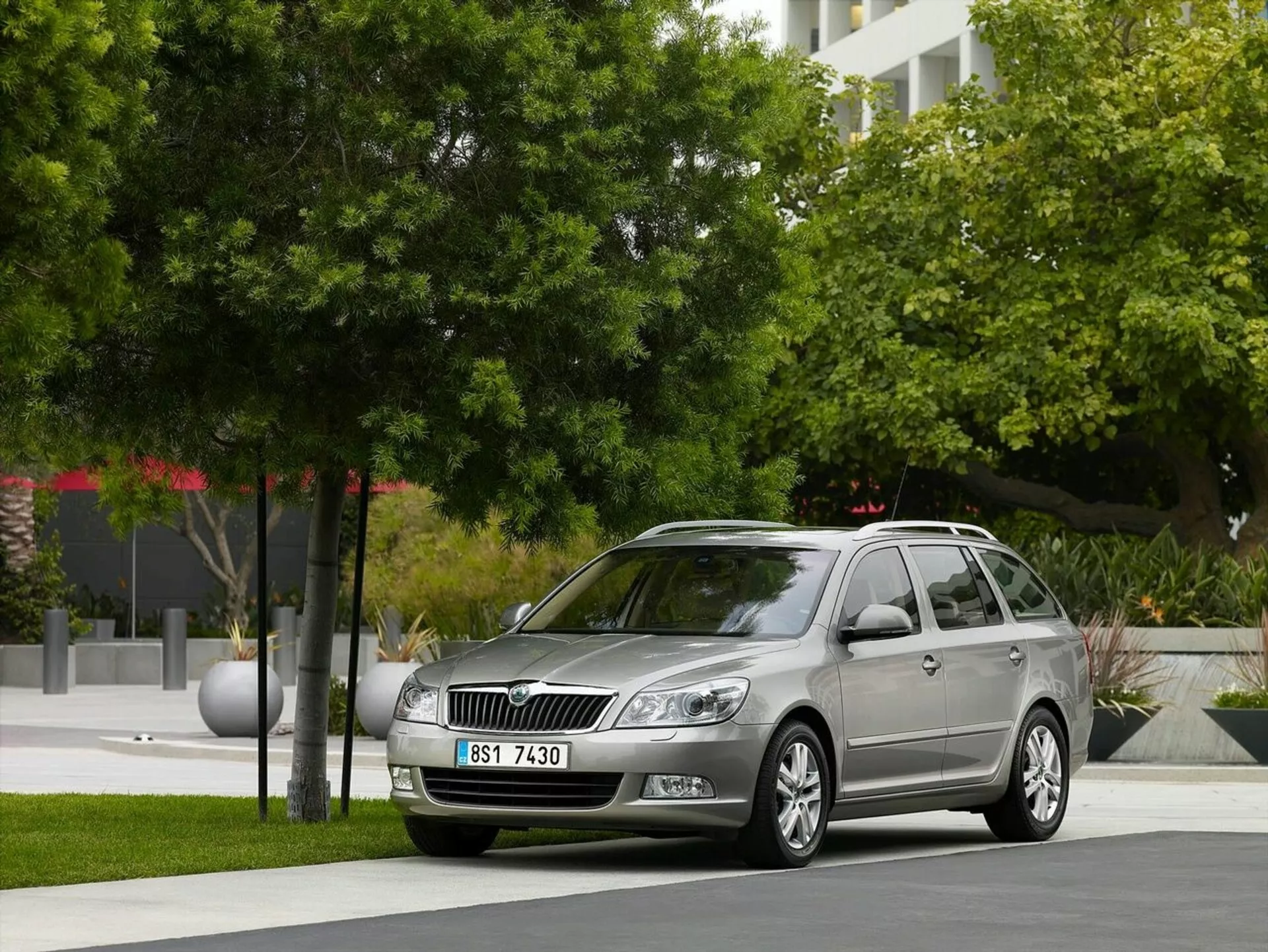 Opel Astra kombi z 2011 roku zaparkowany w miejskim otoczeniu z zielenią i nowoczesnymi budynkami w tle, prezentujący przedni grill samochodu, reflektory i elegancki profil boczny wraz z felgami aluminiowymi i relingami dachowymi