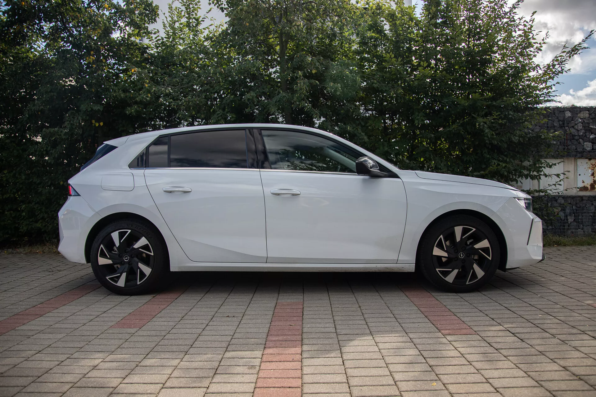 Biały sedan Opel Astra 2022 zaparkowany na zewnątrz, pokazany z profilu bocznego, podkreślający jego pełną długość, aerodynamiczne linie projektowe, koła ze stopu z wieloramiennej konstrukcji i lekko przyciemniane okna w miejskim otoczeniu