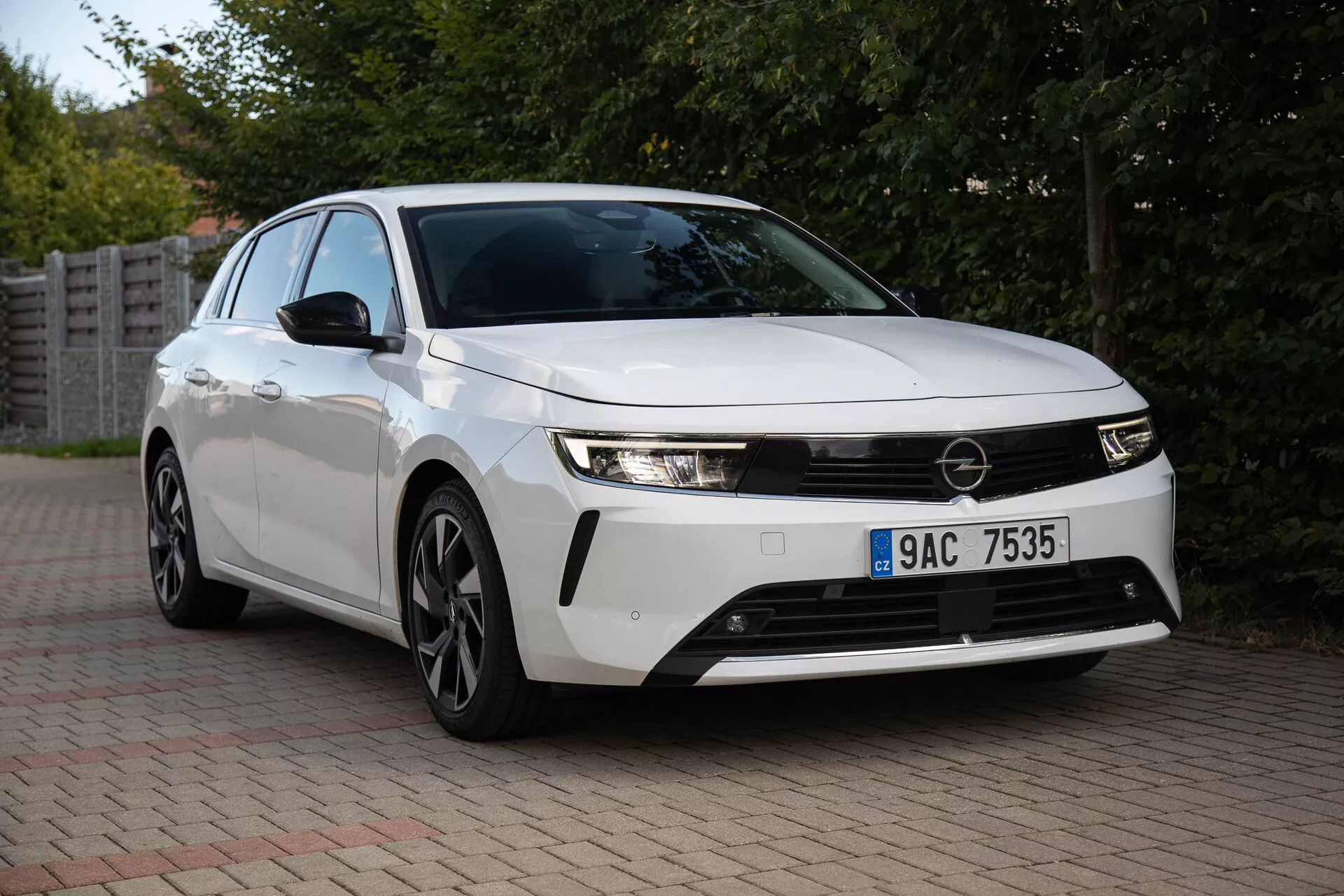 Opel Astra 2022 uchwycony w podmiejskim otoczeniu, pokazujący swoje eleganckie reflektory, wyraźną przednią grillę, aerodynamiczny design w tym koła ze stopu o wielu szprychach, i innowacyjną technologię z widoku z przodu pod trzy czwarte