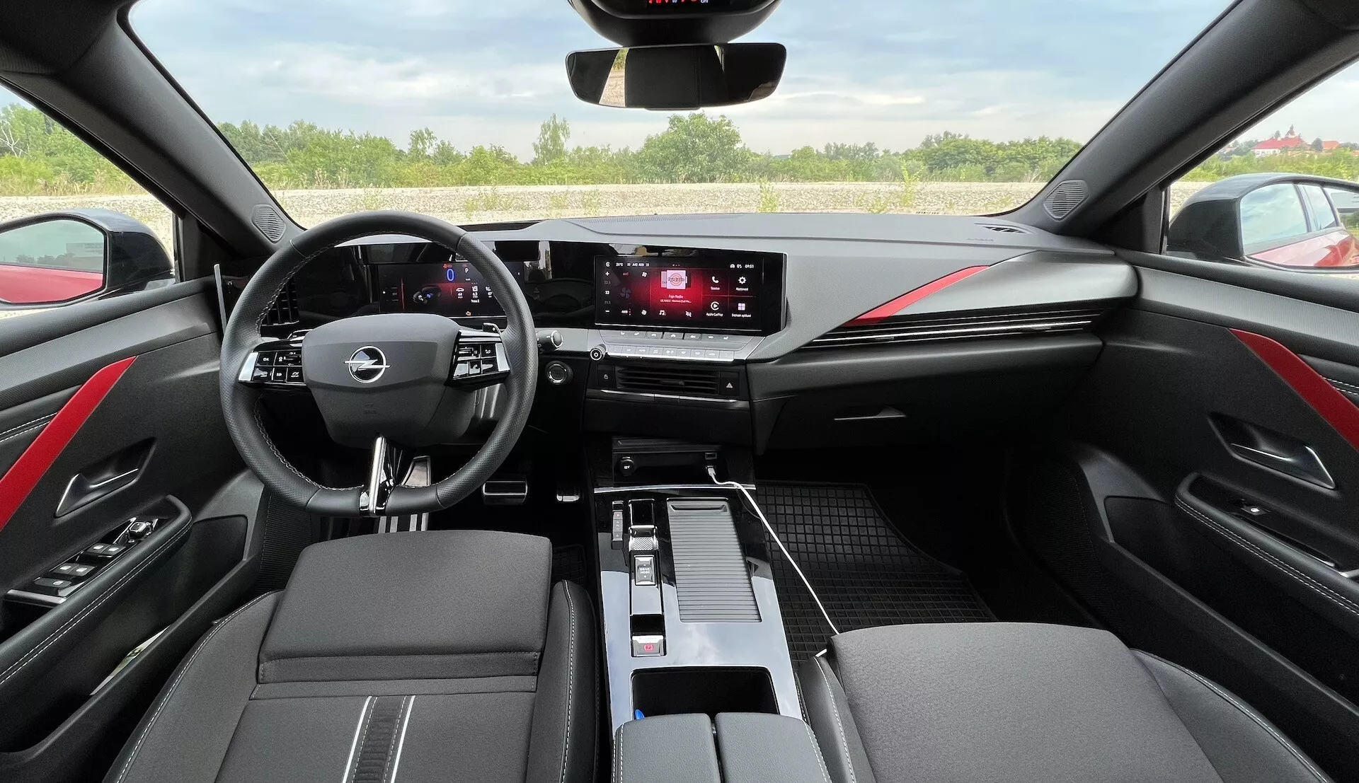 Wnętrze Opel Astra 2022 pokazujące wielofunkcyjną kierownicę z trzema ramionami, cyfrowy zegar, system infotainment z ekranem dotykowym, eleganckie przyciski klimatyzacji, dwukolorowe wykończenia z czarnej skóry z czerwonymi szwami oraz nowoczesny design kabiny z perspektywy kierowcy