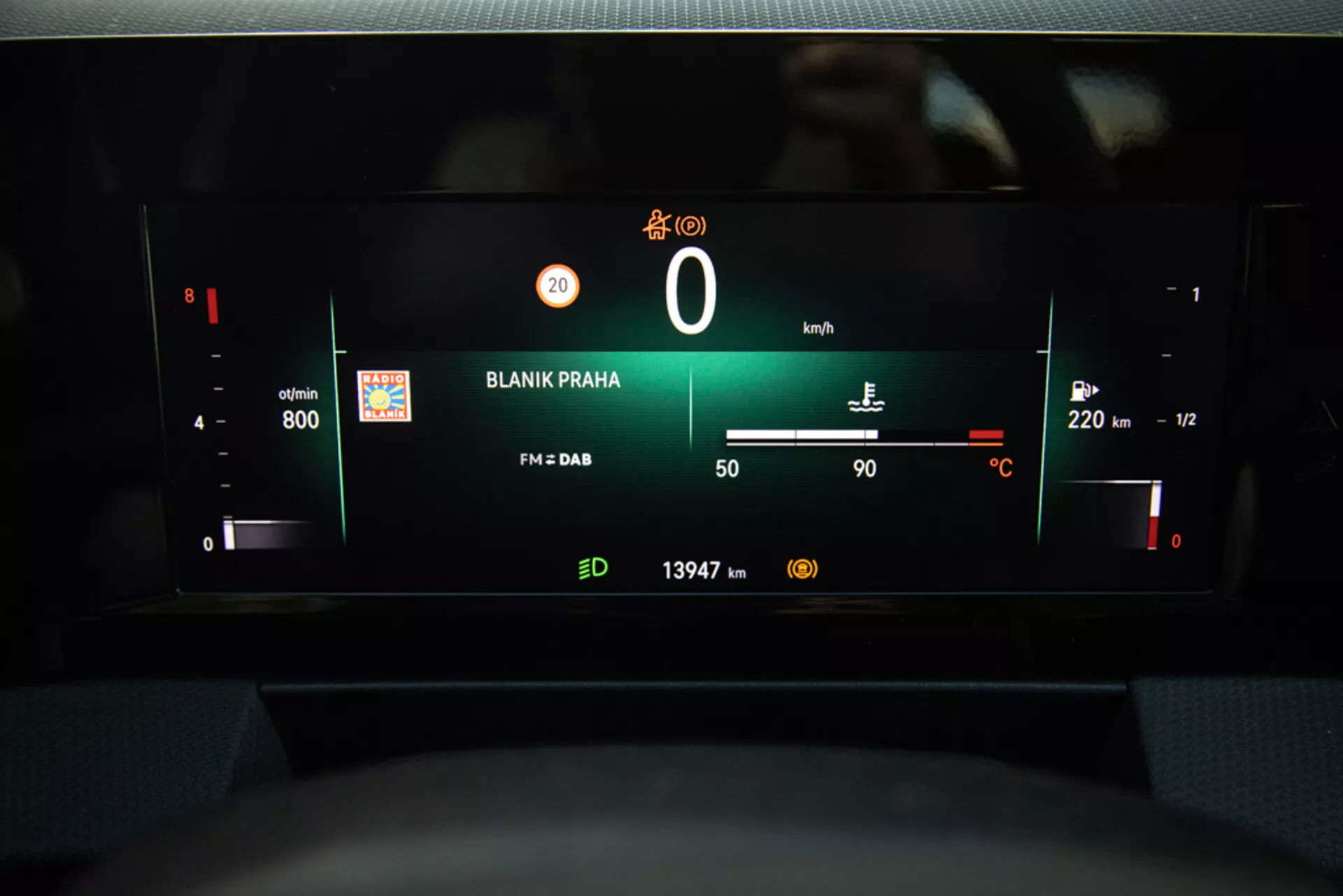 Widok cyfrowego deski rozdzielczej Opel Astra 2022, podkreślający zespół wskaźników z obrotomierzem, poziomem baterii, prędkościomierzem, wskaźnikiem paliwa, temperaturą zewnętrzną, stacją radiową 'BLANIK PRAHA' i odczytem licznika kilometrów z perspektywy miejsca kierowcy.