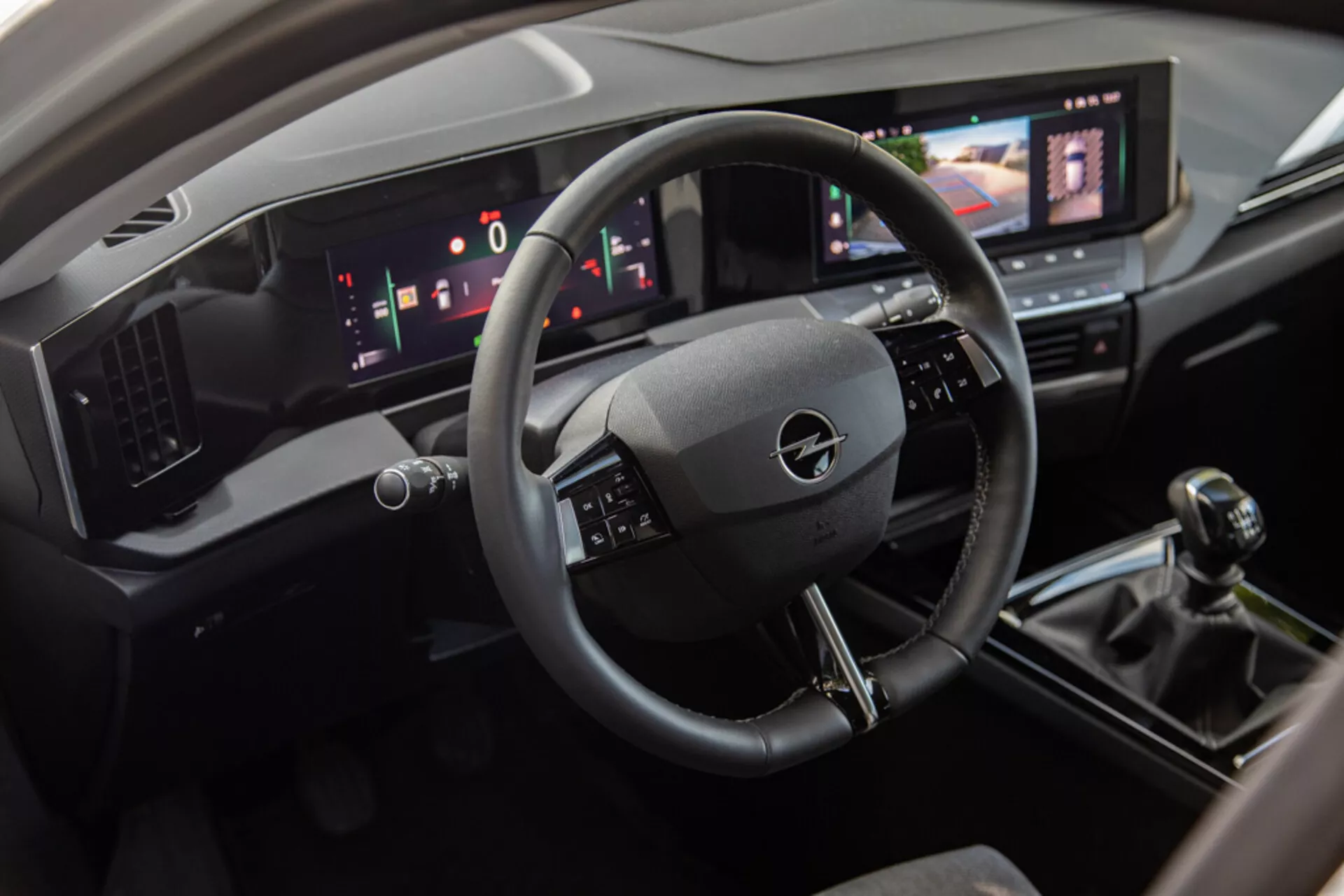 Wnętrze Opel Astra 2022 prezentujące obszar kierowcy z kierownicą o trzech ramionach, cyfrowym deskiem rozdzielczym i centralną konsolą, w tym systemem infotainment z ekranem dotykowym, manualną skrzynią biegów i nowoczesnymi kontrolami klimatyzacji