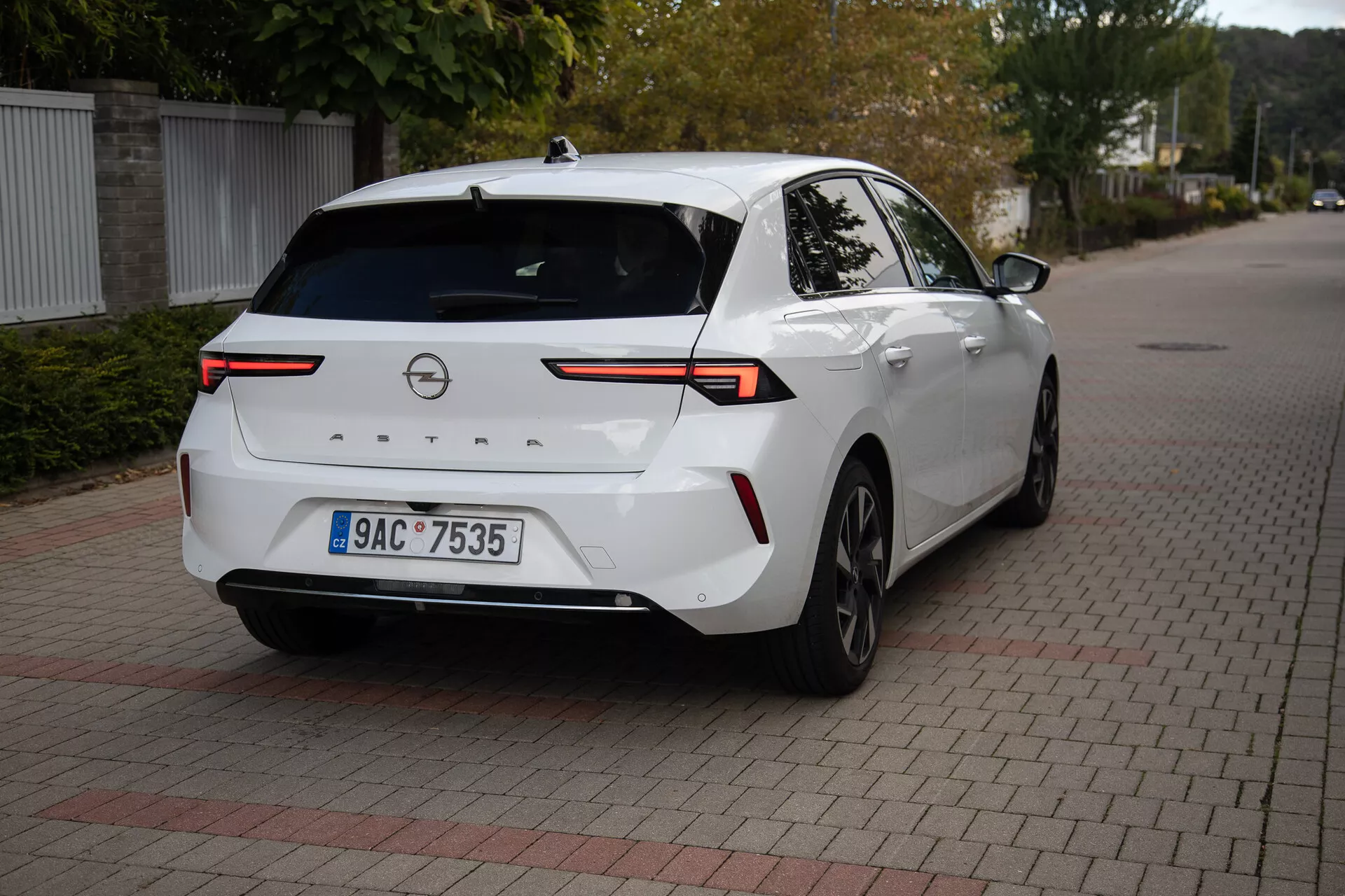 Nowoczesny sedan Opel Astra 2022 w kolorze białym, zaparkowany na nawierzchni z zazębiających się cegieł, prezentujący kąt tylny trzy czwarte z akcentem na zaawansowane technologicznie tylne światła LED, kamerę cofania, czujniki parkowania, częściowy profil boczny w tym prawe tylne koło i drzwi, połączone przez poziomy pas świetlny na klapie bagażnika, wyposażony w antenę płetwy rekina dla ulepszonej łączności, odpowiedni do ilustrowania tylnego designu pojazdu i nowoczesnej technologii samochodowej
