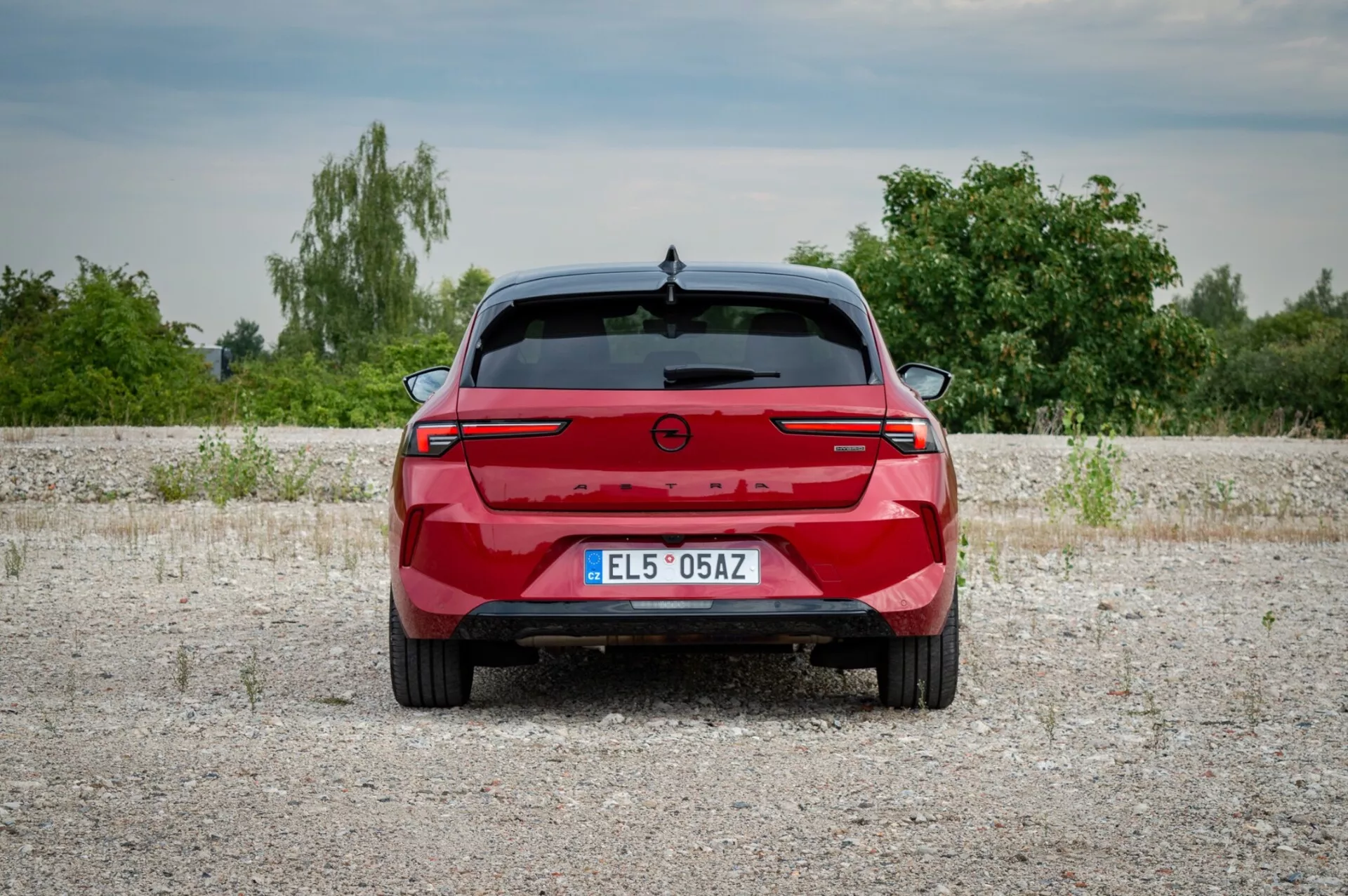 Opel Astra 2022 sfotografowany od tyłu na żwirowej nawierzchni prezentujący tylne światła z łączącą je belką świetlną, czujniki parkowania i brak widocznych portów wydechowych wskazujących na potencjalny model elektryczny lub hybrydowy, na tle roślinności i lekko zachmurzonego nieba.