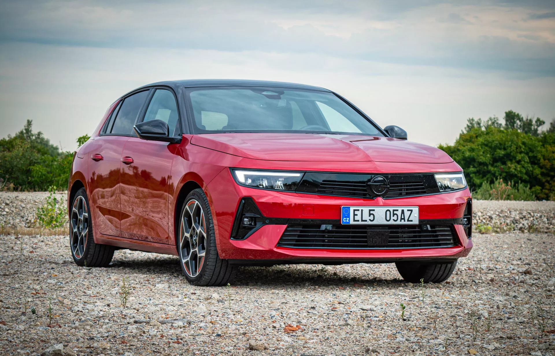 Opel Astra 2022 w żywym czerwonym kolorze, pokazujący nowoczesny design z kątowymi reflektorami LED, wyrazistą przednią kratą, sportowym zderzakiem przednim i felgami ze stopu o wielu szprychach, prezentowany w naturalnym otoczeniu na zewnątrz z widoku trzech czwartych przednich, podkreślający aerodynamiczny profil i luksusowe cechy pojazdu