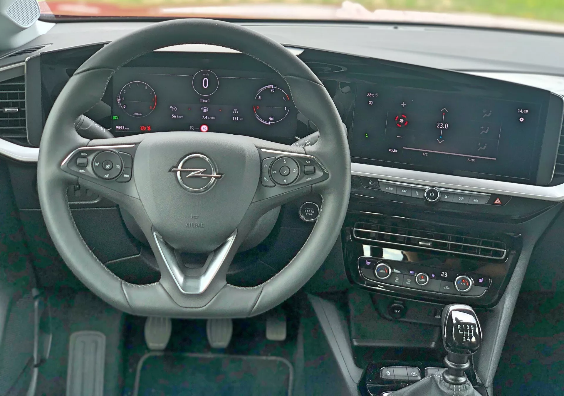 Widok wnętrza Opel Mokka 2021 z perspektywy siedzenia kierowcy, pokazujący trójramienną kierownicę z metalowym emblematem marki, przełączniki na kierownicy do sterowania audio, telefonem i tempomatem, łopatki do zmiany biegów, cyfrowy zestaw wskaźników pokazujący prędkość, obroty, poziom paliwa i temperaturę silnika, wyraźny system informacyjno-rozrywkowy z szerokim poziomym ekranem, dotykowe sterowanie HVAC, dźwignię zmiany biegów z osłoną ze skóry, polerowane elementy wykończeniowe i przycisk start-stop.