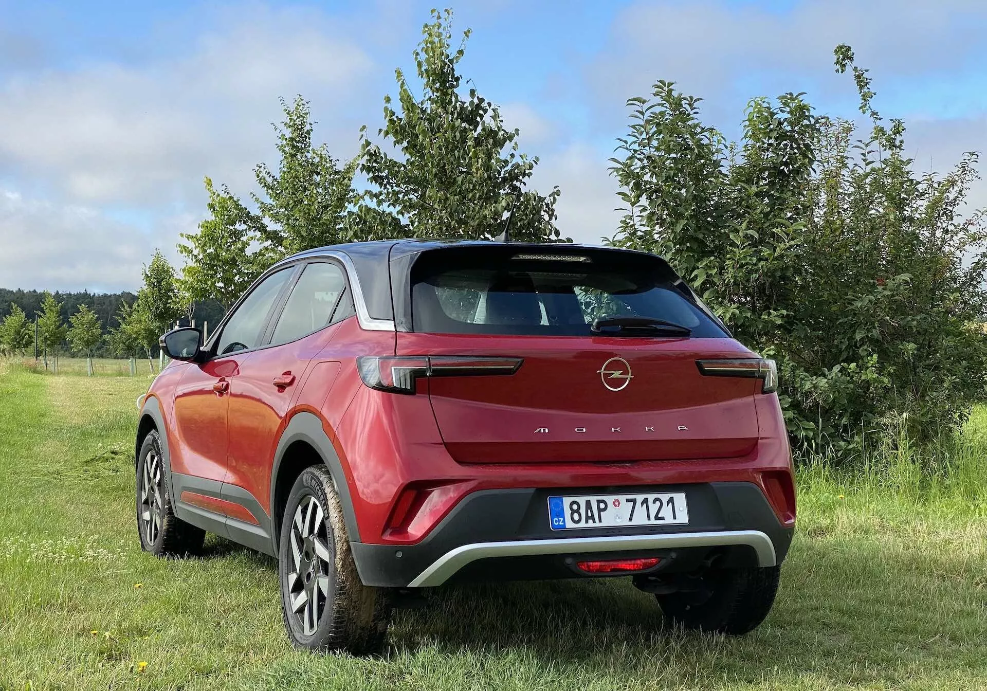 Kolorowa fotografia przedstawiająca tylny trzyczwarty widok nowoczesnego crossovera SUV Opel Mokka 2021 w czerwono-czarnym kolorze, zaparkowanego na zewnątrz na trawiastym terenie z drzewami lub krzewami w tle. Elementy designu pojazdu obejmują aerodynamiczny tylny spoiler, kątowe tylne światła LED, wysoko zamontowaną lampę stopu, dwukolorowe nadwozie, duże aluminiowe felgi, wpuszczane klamki drzwi i czarne plastikowe listwy. Zdjęcie zostało zrobione z niskiego kąta, ukazując wysokość i prześwit SUV-a, z częściowo pochmurnym niebem wskazującym na zmienne warunki pogodowe.
