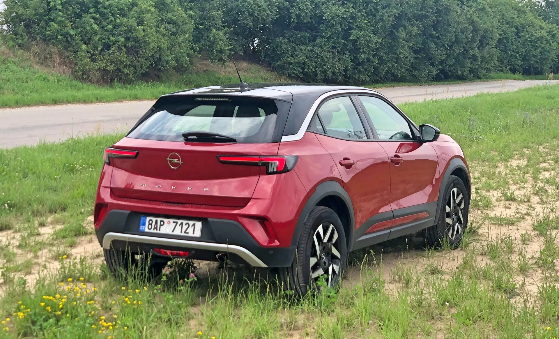 Nowoczesny crossover SUV Opel Mokka 2021 widziany z tylnej trójczwartej perspektywy, podkreślający tylny spojler, stromo pochyloną tylną szybę, widoczny znak firmowy i eleganckie tylne światła. Ustawiony na trawiastej powierzchni, pokazując swój potencjał do lekkiej jazdy terenowej. Profil boczny ujawnia rzeźbione linie nadwozia, czarne osłony i stylowe wieloszprychowe koła. Zintegrowana tylna wycieraczka, czujniki parkowania i kontrastowa osłona podwozia sugerują aerodynamikę, praktyczność i wytrzymałość.
