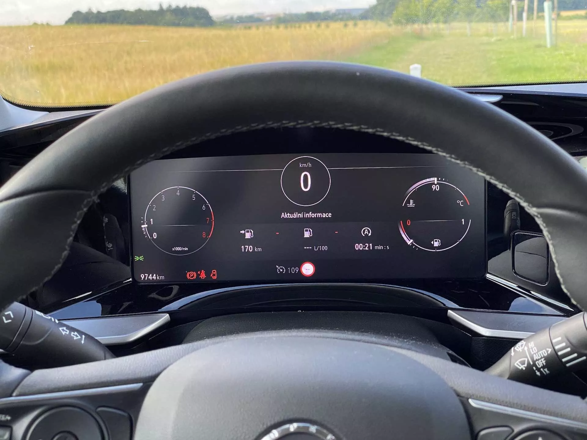 Widok wnętrza Opel Mokka 1.2 DI Turbo 2021 z perspektywy kierowcy za kierownicą. Obraz podkreśla cyfrowy zestaw wskaźników za kierownicą, który obejmuje prędkościomierz, obrotomierz i panel informacyjny wyświetlający temperaturę zewnętrzną (8°C), zegar, pozycję biegu ('P' dla parkowania) i szczegóły komputera pokładowego. Kierownica ma widoczne przyciski do mediów i jest częściowo widoczna. Oświetlenie otoczenia sugeruje dzień z widokiem na wiejski krajobraz przez przednią szybę. Pojazd posiada elektroniczny wyświetlacz deski rozdzielczej i zaawansowane funkcje technologiczne.