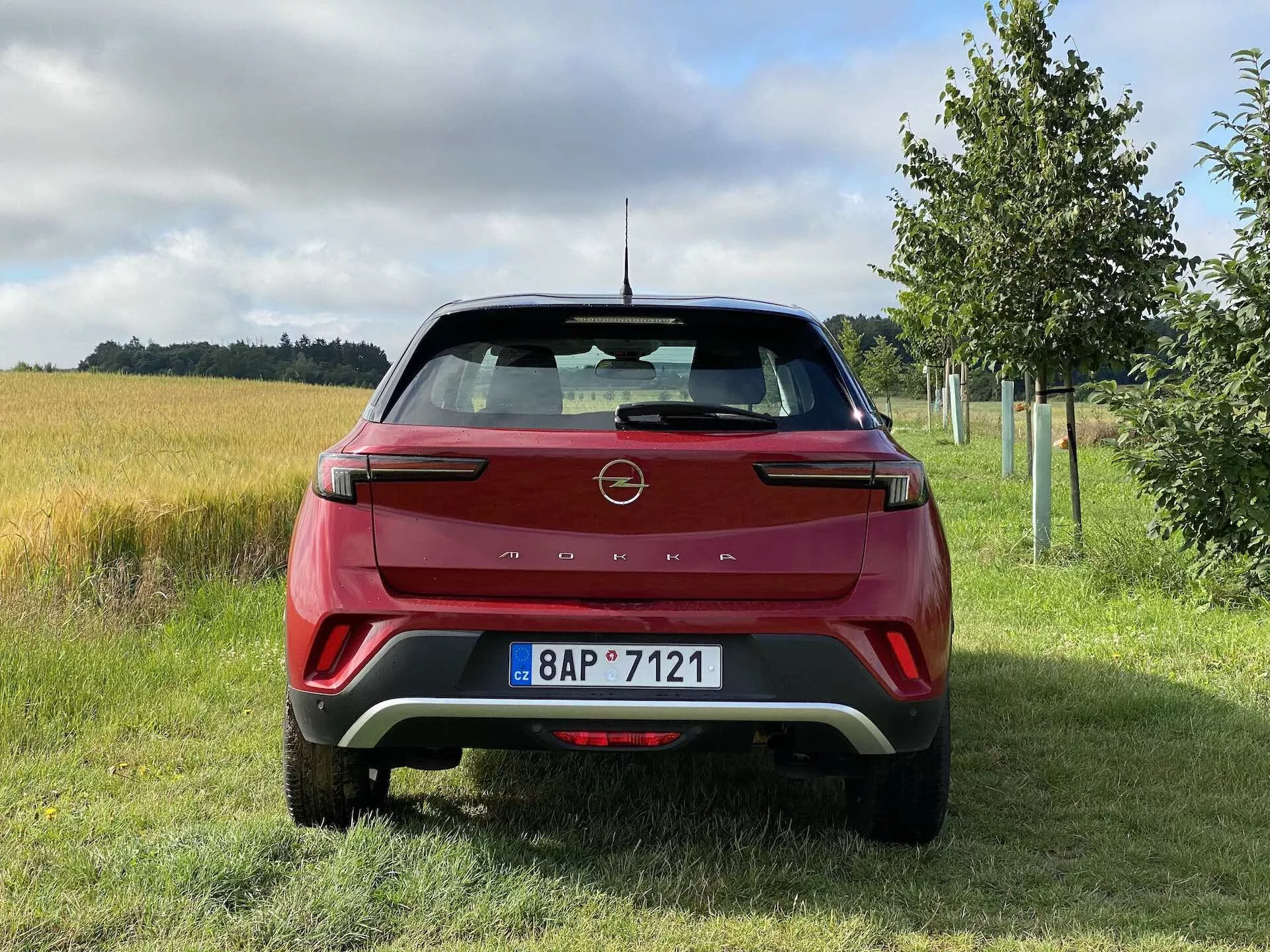 Tylny widok czerwonego Opel Mokka 1.2 DI Turbo 2021, prezentującego nowoczesny design z ostrymi liniami i emblematem producenta na tylnej klapie, charakterystycznymi klastrowymi światłami tylnymi LED, tylnym zderzakiem z kontrastowymi wstawkami, czujnikami parkowania, zintegrowanymi odblaskami, wycieraczką tylnej szyby, anteną typu 'płetwa rekina', tylnym spojlerem i tablicą rejestracyjną w wiejskim otoczeniu z trawą, polem uprawnym i drzewami.