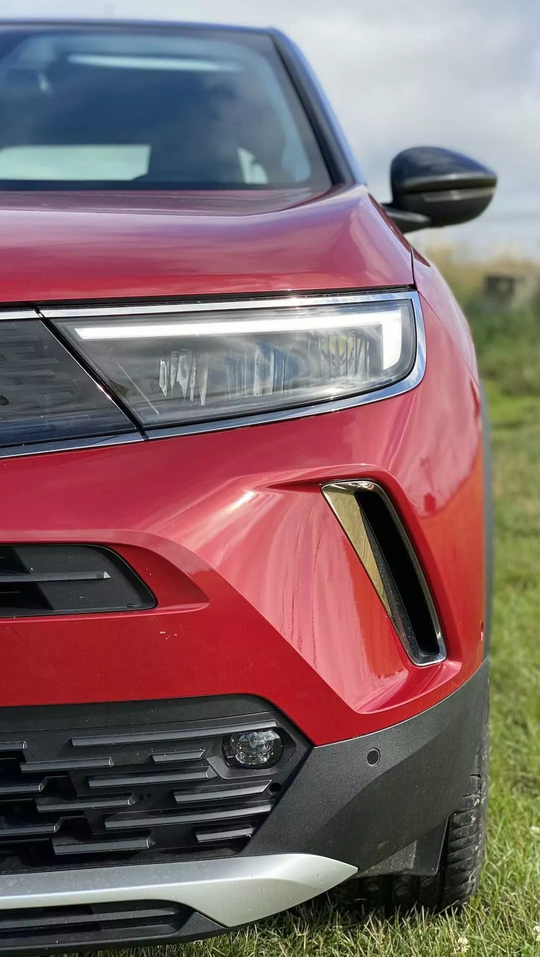 Zbliżony, skośny widok na przednią lewą stronę czerwonego Opel Mokka 2021. Na zdjęciu widoczny reflektor z oświetleniem LED lub HID, część grilla z poziomymi listwami, obudowa światła przeciwmgielnego z czarno-srebrnym obramowaniem, żywy czerwony lakier karoserii z ostrymi, kanciastymi konturami, opływowe lusterko boczne i częściowo widoczna maska i błotnik. Samochód jest zaparkowany na trawie z rozmytym tłem.