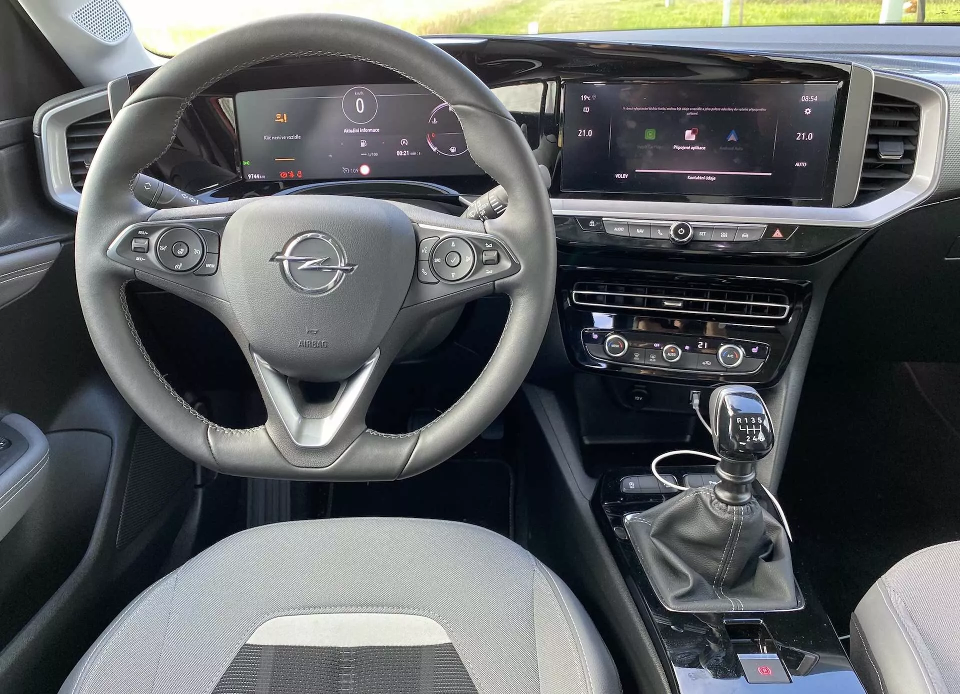 Wnętrze Opel Mokka 2021 z perspektywy przedniego pasażera, z trójramienną kierownicą z zamontowanymi przyciskami, w pełni cyfrowym panelem instrumentów, centralnym ekranem dotykowym systemu infotainment z kontrolą klimatu i nawigacją oraz manualną dźwignią zmiany biegów. Układ obejmuje podwójne systemy kontroli i dodatkowe przyciski do trybów jazdy, z tapicerką z tkaniny.