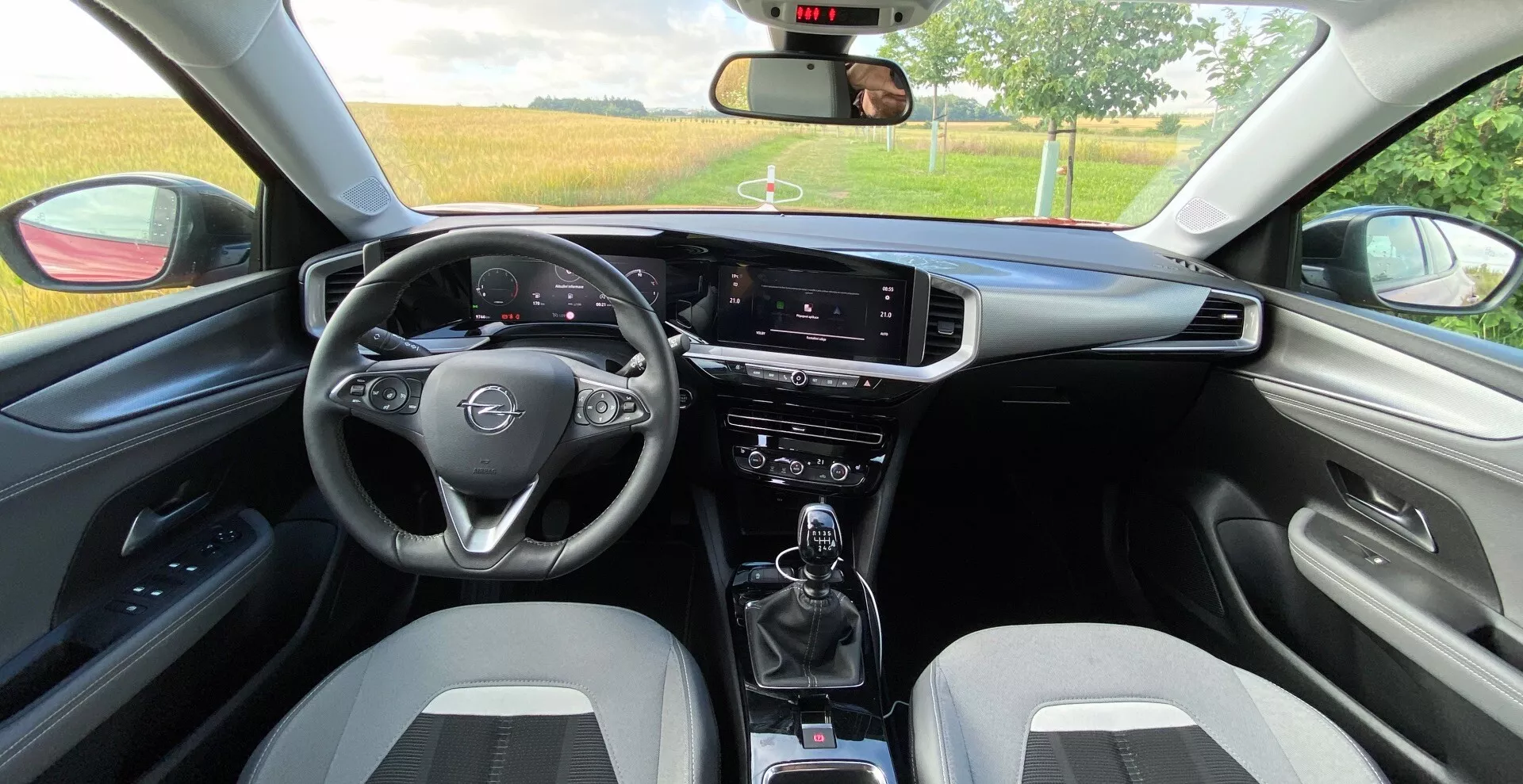 Widok wnętrza z tylnego siedzenia samochodu Opel Mokka 2021 1.2 DI Turbo. Zawiera trójramienną kierownicę wielofunkcyjną, cyfrowy wyświetlacz prędkościomierza i obrotomierza za kierownicą, pionowy dotykowy ekran infotainment, przełączniki klimatyzacji, manualną skrzynię biegów z uchwytami na kubki, sportowe fotele przednie dwukolorowe, dopasowane panele drzwi z podłokietnikiem, widoczne lusterko wsteczne, częściowo opuszczone szyby i dodatkowe dźwignie do wycieraczek i kierunkowskazów.