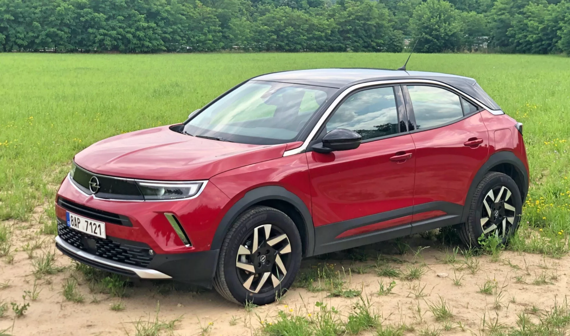 Obraz czerwonego SUV-a Opel Mokka 2021 zaparkowanego na trawiastej powierzchni z polem w tle. Widok z trzech czwartych z przodu podkreśla kratkę, potencjalnie wskazującą na zaawansowaną technologię układu napędowego, eleganckie diodowe światła do jazdy dziennej, światła przeciwmgielne i unikalne dwukolorowe aluminiowe felgi. Widoczne są również lusterka i klamki drzwi w kolorze nadwozia, dynamiczna linia ramienia, ochronna dolna listwa czarna i chromowane ramki okien, prezentujące nowoczesne i luksusowe cechy projektowe pojazdu.