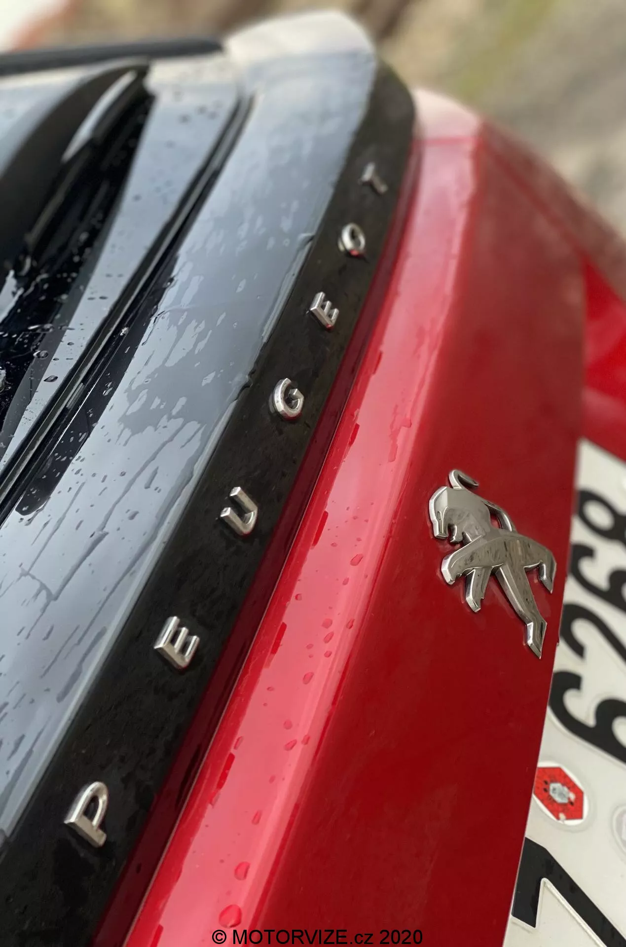 Zbliżenie detalem tylnego emblemat i oznaczenia modelu Peugeot 2008 2020 (1.2 PureTech 100 Active). Zdjęcie jest wykonane pod nietypowym kątem diagonalnym, pokazując logo Peugeot z chromowanym wykończeniem na tle jaskrawej czerwonej powierzchni nadwozia. Chromowane litery rozmieszczone po lewej stronie loga tworzą napis 'PEUGEOT'. Powyżej nazwy widoczna jest błyszcząca czarna powierzchnia z kroplami wody, co może wskazywać na niedawny deszcz lub mycie samochodu. Rozmazana część tablicy rejestracyjnej jest widoczna poniżej i po prawej stronie. Płytka głębia ostrości uwydatnia emblemat i nazwę, podczas gdy reszta elementów jest zamazana dla artystycznego efektu.
