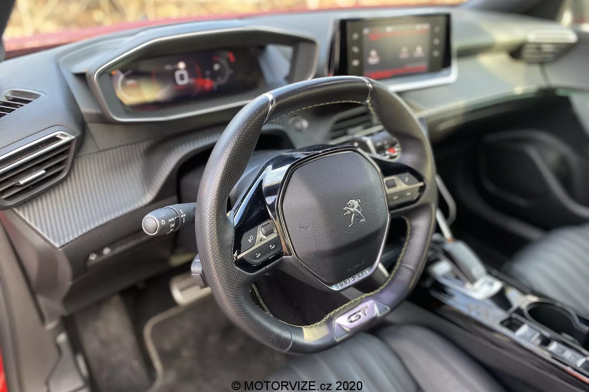 Wnętrze Peugeot 2008 1.2 PureTech 100 Active z 2020 roku z perspektywy przedniego pasażera, skupione na kokpicie kierowcy. Obraz pokazuje płaski dół kierownicy z wieloma przyciskami sterującymi i logo lwa Peugeota, cyfrowy zestaw wskaźników, system infotainment z ekranem dotykowym, centralną konsolę automatycznej skrzyni biegów, przycisk rozrusznika/zatrzymania, wyloty powietrza z chromowanymi akcentami oraz mieszankę miękkich materiałów, w tym skóry i tekstur przypominających włókno węglowe.