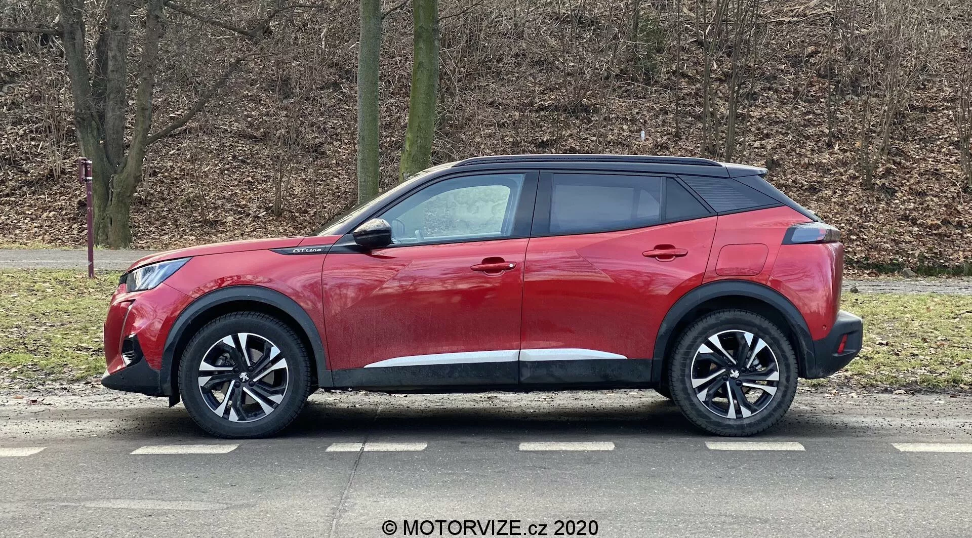 Czerwony SUV Peugeot 2008 2020 1.2 PureTech 100 Active o nowoczesnym i eleganckim designie, pochylona linia dachu, wysoka linia okien z wąskim zwieńczeniem, aerodynamiczne klamki drzwi, lusterka boczne z wbudowanymi kierunkowskazami, duże wieloszprychowe felgi aluminiowe, niskoprofilowe opony, czarne plastikowe osłony nadające wytrzymały wygląd, połyskujący czerwony lakier oraz przyciemniane tylne szyby, uchwycony z profilu ustawiony na zewnątrz z tłem z bezlistnymi drzewami i trawnikiem.