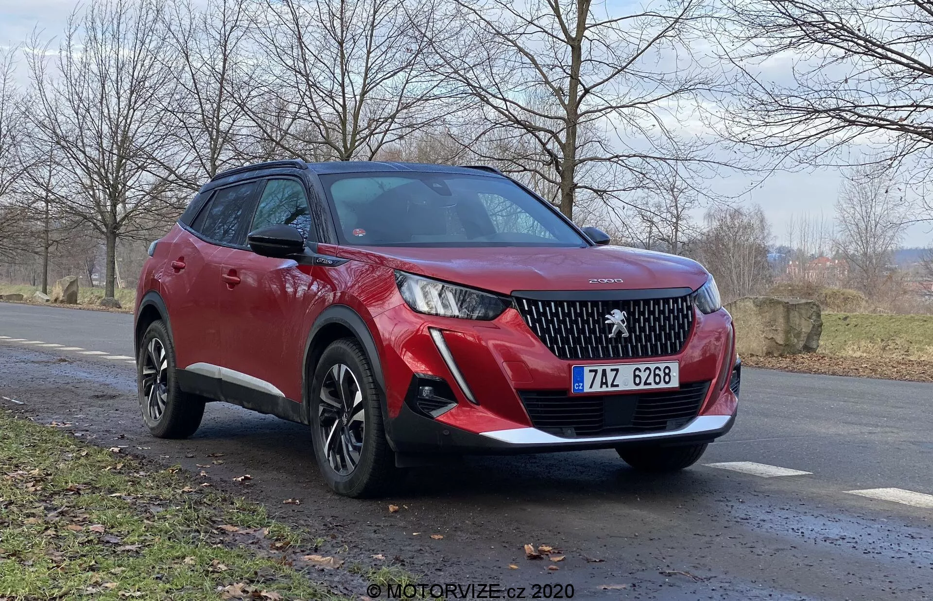 Czerwony SUV Peugeot 2008 2020 (Peugeot-2008-1-2-Puretech-100-Active) z widokiem z przodu trzech czwartych, podkreślający przedni grill z wyraźnym emblematem marki, eleganckimi reflektorami LED, przednimi czujnikami parkowania i światłami przeciwmgielnymi. Widok boczny ujawnia wieloramienne felgi aluminiowe, wyższą wysokość zawieszenia, dynamiczne linie nadwozia, zintegrowane kierunkowskazy na lusterkach bocznych i czarne plastikowe osłony. Ustawiony pod lekkim kątem z boku drogi z naturalnym, wiejskim krajobrazem w tle, wskazującym na późną jesień lub wczesną wiosnę. Niski kąt kamery dla potężnego, imponującego widoku.