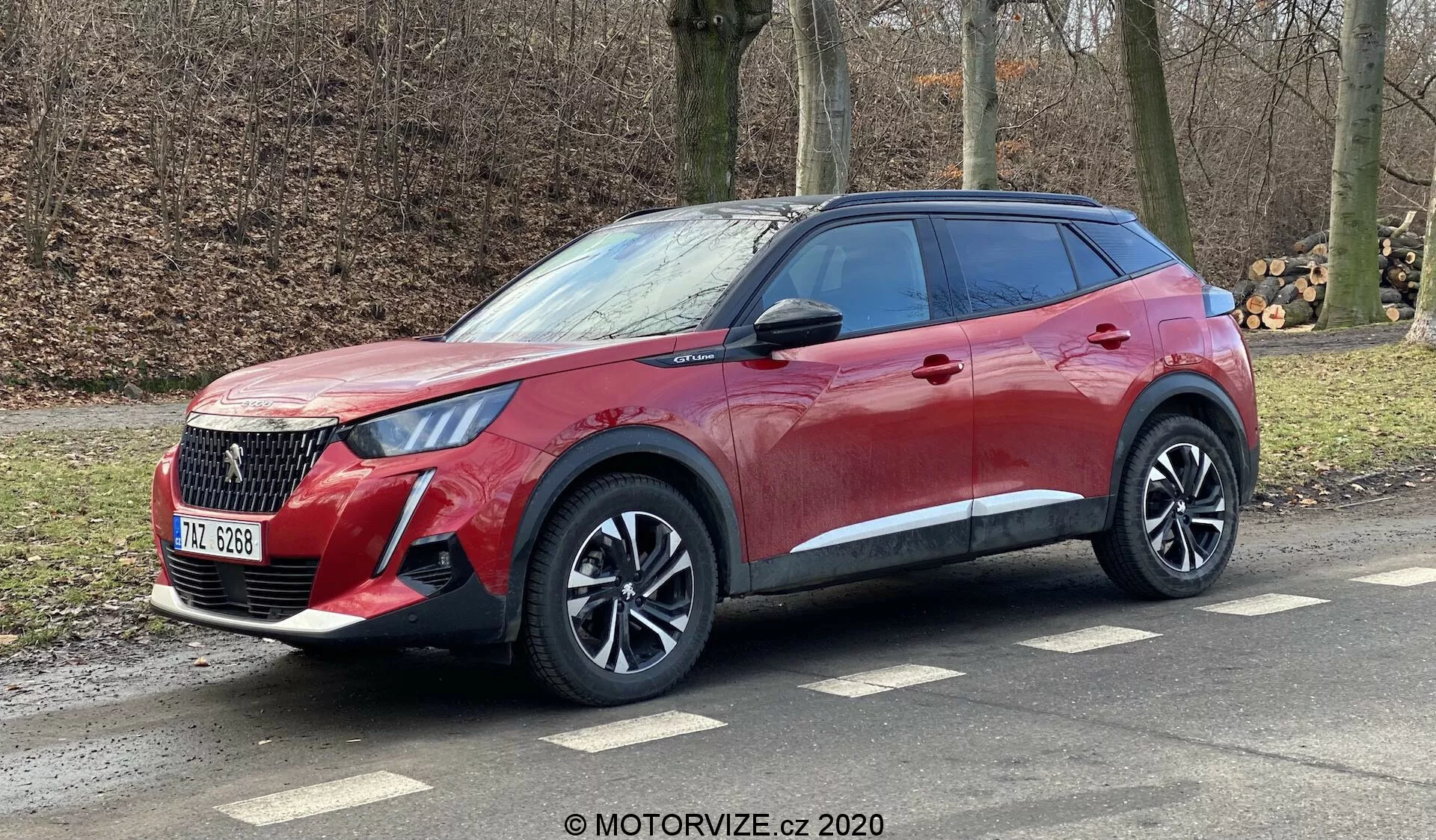 Perspektywa boczno-przednia nowoczesnego crossover SUV Peugeot 2008 1.2 PureTech 100 Active 2020, podkreślającego jego wyrazistą kratkę, eleganckie reflektory, aluminiowe felgi i dynamiczne linie nadwozia. Pojazd, ozdobiony czerwonym metalizowanym wykończeniem z przyciemnianymi tylnymi szybami, jest pokazany podczas parkowania na bocznej drodze z widocznymi relingami dachowymi, sugerując zdolność do jazdy miejskiej i lekkiej terenowej. Źródło zdjęcia: MOTORVIZE.cz 2020.