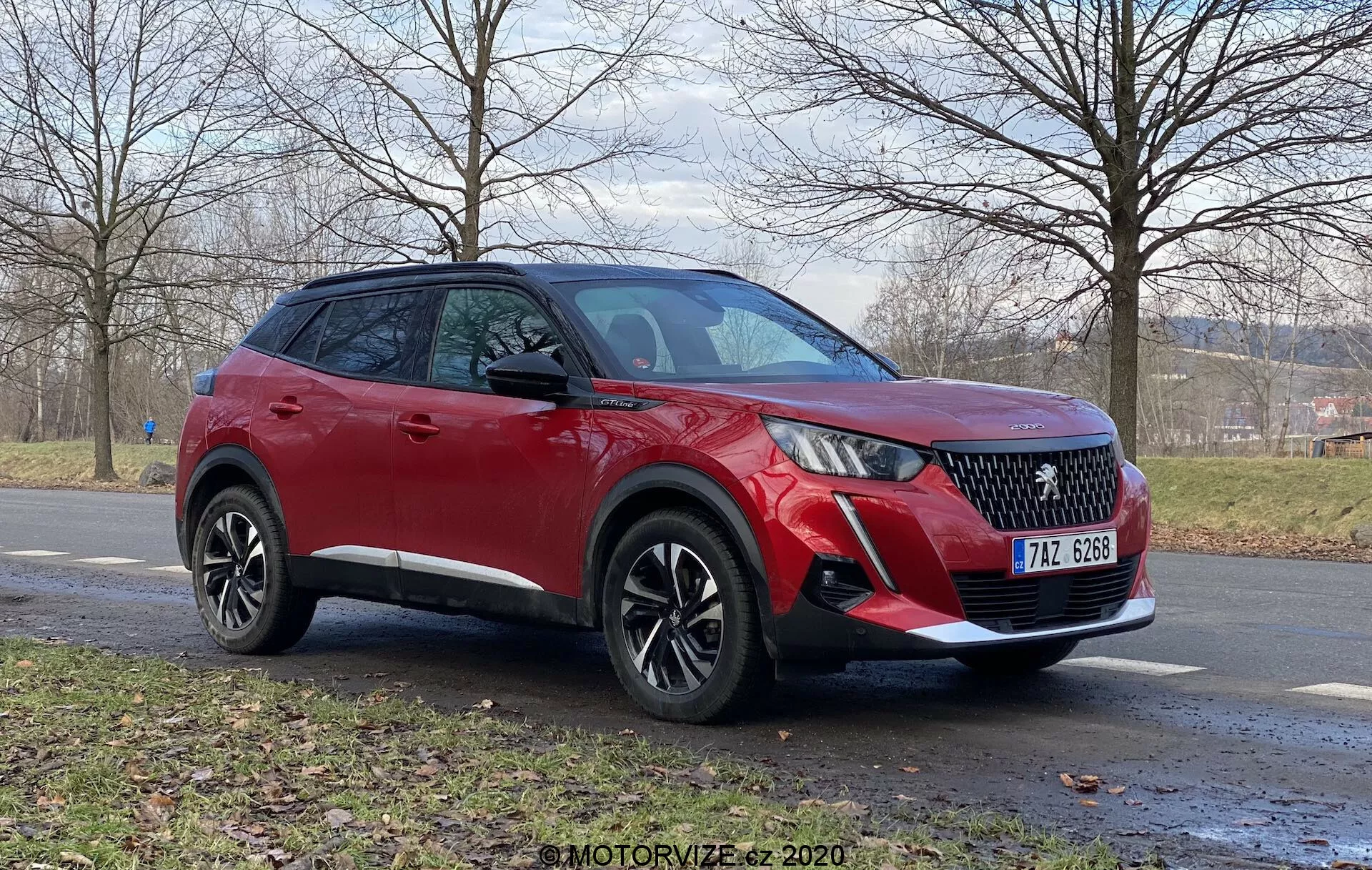 Widok trzech czwartych przodu czerwonego Peugeot 2008 2020 (1.2 PureTech 100 Active) crossover SUV na nawierzchni asfaltowej. Widoczne cechy obejmują LED-owe światła do jazdy dziennej, kratkę w diamentowy wzór, przedni zderzak z światłami przeciwmgielnymi i możliwymi czujnikami parkingowymi, wieloramienne felgi aluminiowe oraz kamerę pod lusterkiem bocznym w kolorze nadwozia, wszystko pod pochmurnym niebem. W tle widać trawiasty obszar i drzewa bez liści, co sugeruje późną jesień lub zimę.