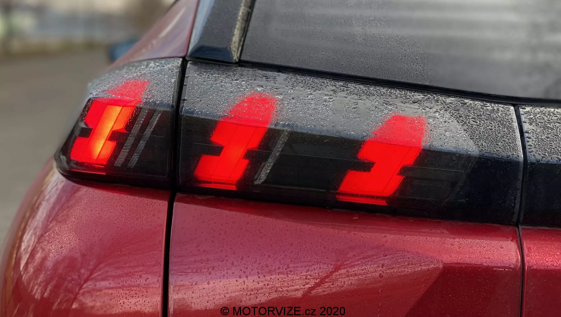 Zbliżenie tylnej części czerwonego Peugeota 2008 2020 (PureTech 100 Active) z naciskiem na zespół tylnych świateł pokazujący zapalone światła hamowania. Fotografia, wykonana z tylnego rogu nieco z boku, podkreśla trzy wyraźne pionowe elementy LED i krople wody na powierzchni, podkreślając nowoczesny design oświetlenia samochodowego.