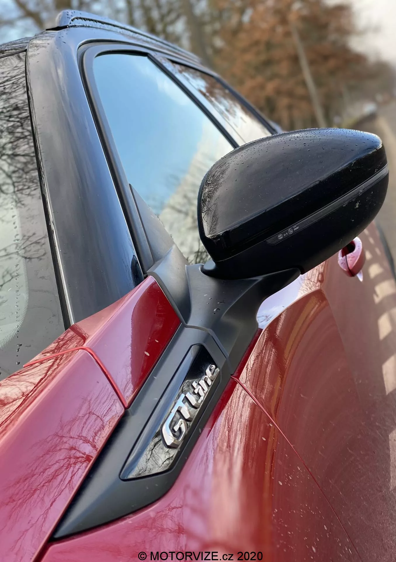 Zbliżenie na bok czerwonego Peugeota 2008 2020, skupiające się na czarnym lusterku bocznym po stronie kierowcy, przymocowane do drzwi z czarnym wspornikiem. Słup A z emblematem 'GT Line' jest częściowo widoczny, pokazując aerodynamiczne linie projektu samochodu i sportowe cechy wykończenia. Znak wodny MOTORVIZE.cz 2020.
