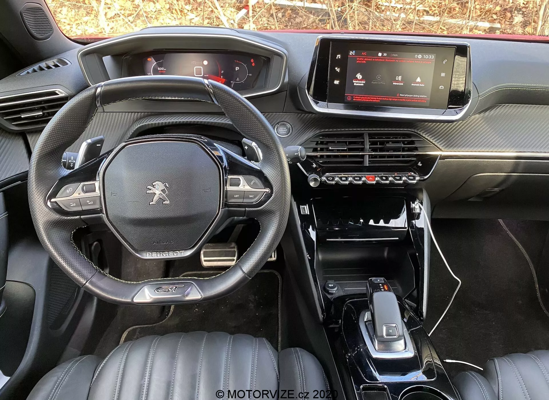 Widok wnętrza Peugeot 2008 1.2 PureTech 100 Active z 2020 roku z tylnego siedzenia, ukazujący deskę rozdzielczą, cyfrowy wyświetlacz infotainment, panel sterowania klimatyzacją, minimalistyczną konsolę środkową z dźwignią zmiany biegów, uchwytami na kubki i dodatkowymi przyciskami sterującymi. Kierownica z płaskim dnem posiada logo Peugeot i wielofunkcyjne przyciski. Przednie siedzenia ze skórzaną tapicerką w pikowany wzór sugerują wyższy poziom wykończenia. Naturalne oświetlenie podkreśla design i materiały fabrycznego modelu.