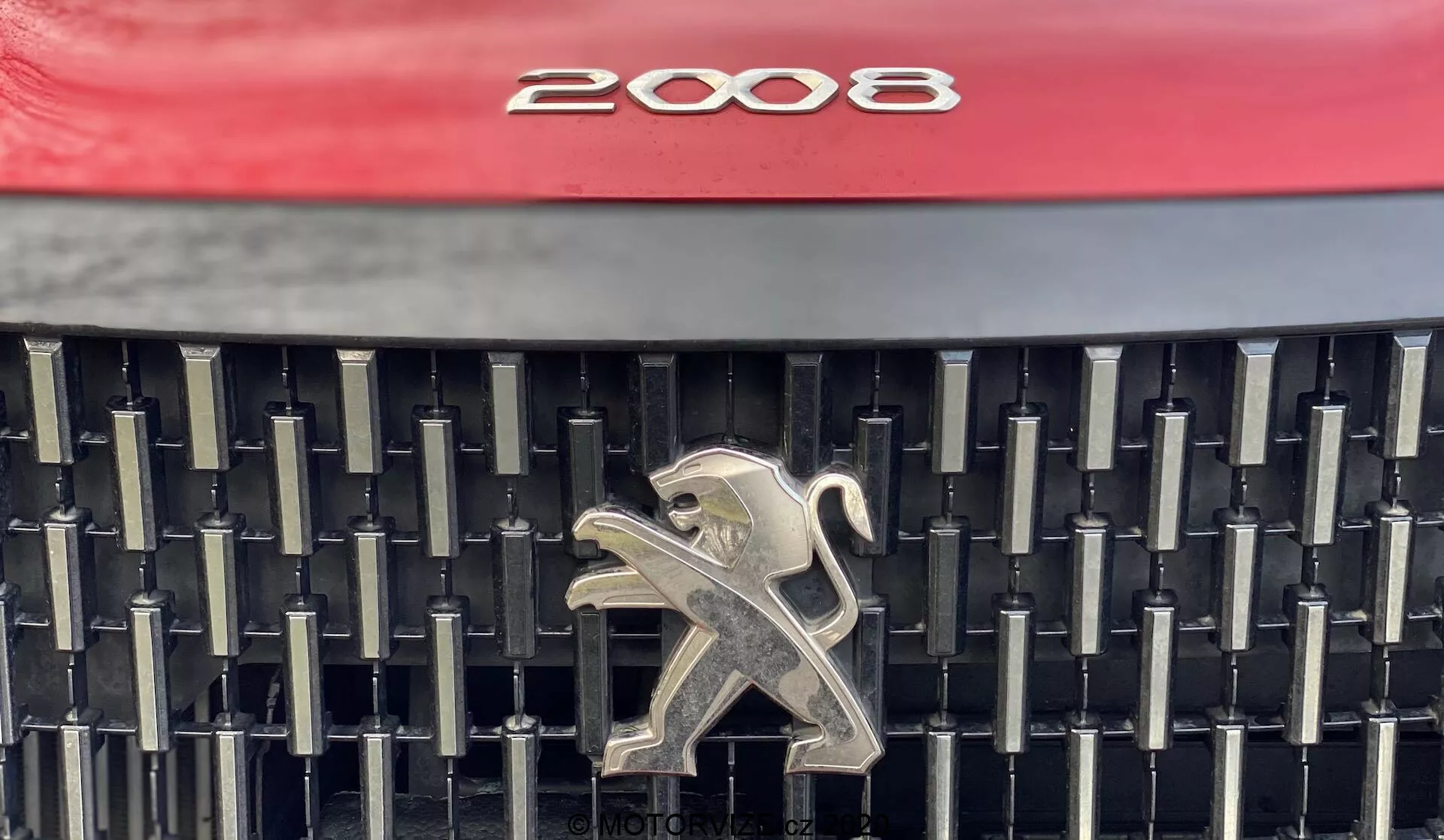Zbliżenie przedniego grilla Peugeot 2008 1.2 PureTech 100 Active 2020, prezentującego charakterystyczny design z jaskrawo czerwonym kapturem, skomplikowaną sieć czarnych pionowych i poziomych listew oraz wypolerowanym srebrnym emblematem lwa pośrodku. Zdjęcie koncentruje się na estetyce i elementach marki, zapewniając wyraźną wizualizację szczegółów grilla.