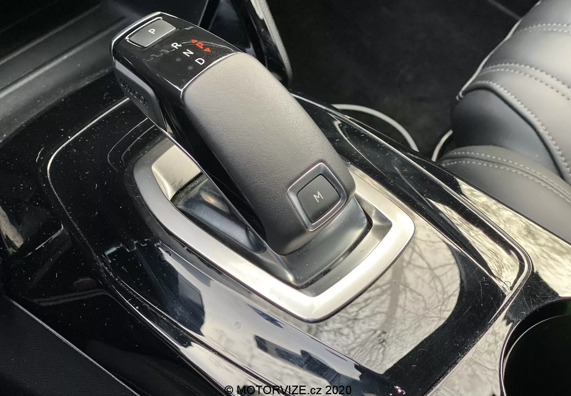 Zbliżenie z bocznej perspektywy wnętrza Peugeot 2008 2020 z elegancką dźwignią zmiany biegów, z automatyczną skrzynią biegów oznaczoną 'P', 'R', 'N', 'D' i 'M' dla trybu manualnego, na błyszczącej czarnej konsoli centralnej z chromowanymi akcentami i skórzanym otoczeniem, co wskazuje na luksusowe wnętrze samochodu.
