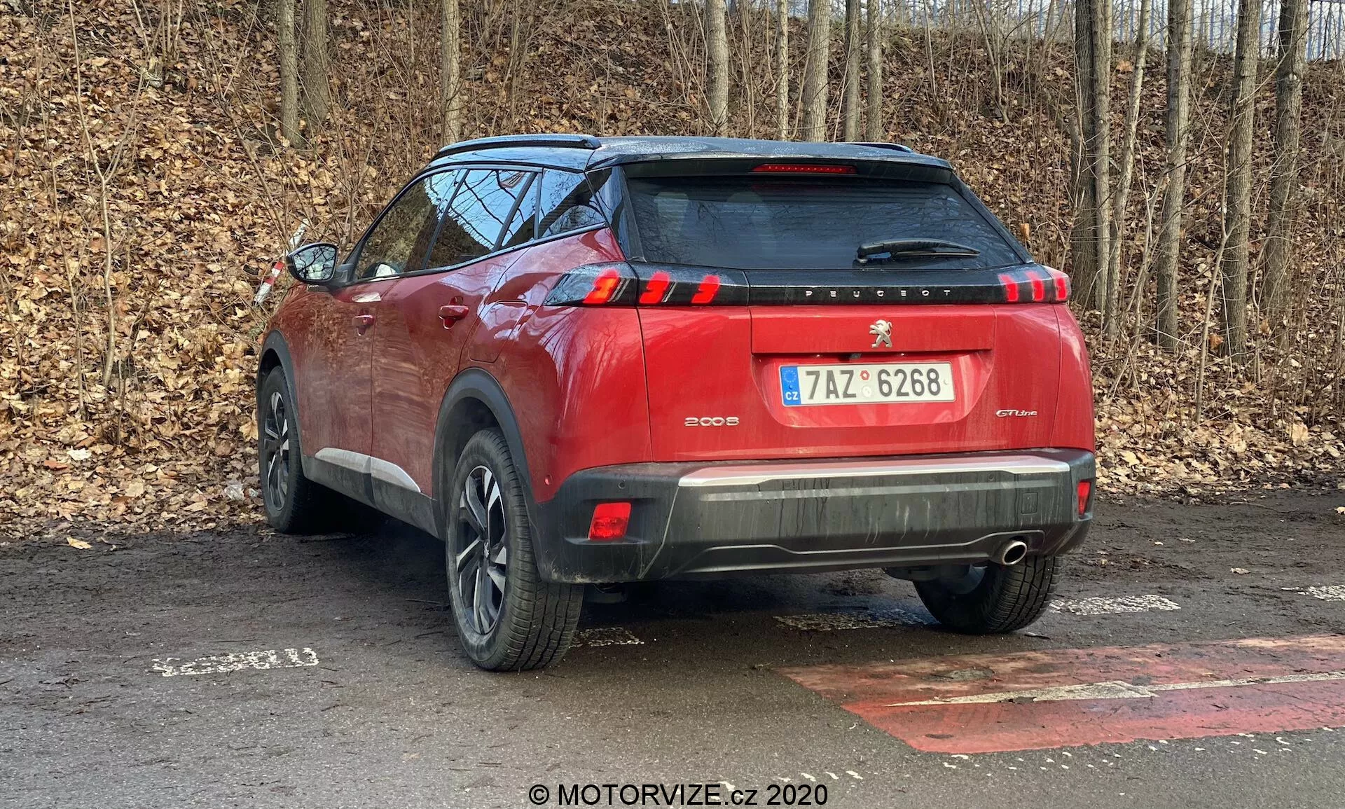 Tylny kąt widzenia czerwonego SUV-a Peugeot 2008 1.2 PureTech 100 Active z 2020 roku zaparkowanego na utwardzonej powierzchni, z tłem pozbawionych liści drzew sugerujących późną jesień lub zimę. Uchwycone z tylnej, trójkwartowej perspektywy, podkreślającej prawą stronę pojazdu. Zdjęcie ukazuje elementy projektu samochodu takie jak tylne światła, tylna szyba, dwukolorowy tylny zderzak z czujnikami parkowania, spojler dachowy, pionowe paski świateł hamowania, częściowo widoczne felgi aluminiowe oraz relingi dachowe. Ukryty wydech może sugerować hybrydowy lub elektryczny układ napędowy. Pojazd zajmuje większość kadru, co sugeruje bliskość fotografa.