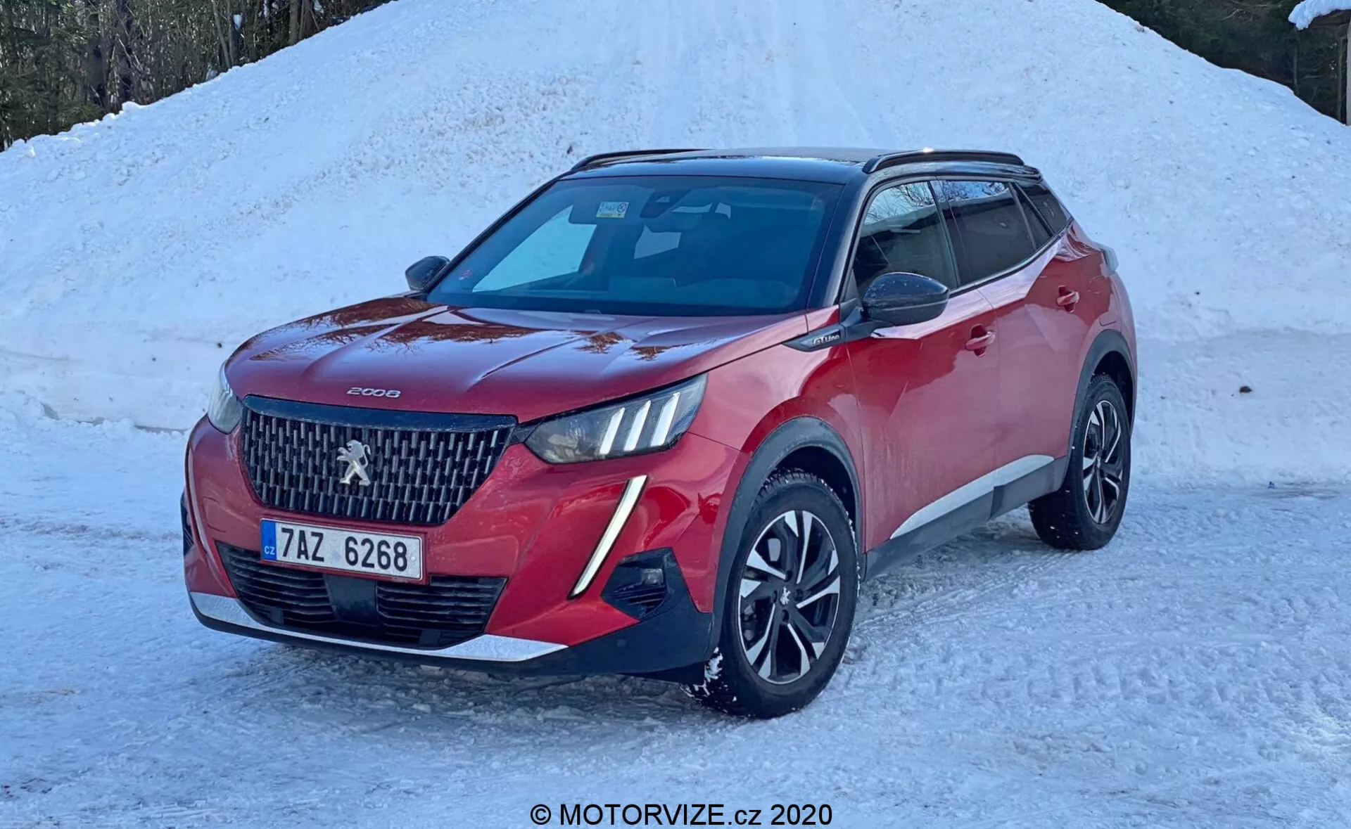 Obraz czerwonego SUV-a, konkretnie Peugeot 2008 2020 (peugeot-2008-1-2-puretech-100-active), zaparkowanego na ziemi pokrytej śniegiem. Nowoczesny pojazd jest pokazany pod kątem trzech czwartych od przodu, podkreślając jego wyraźny grill z dużym centralnym logo i wzorem w diamenty. Widoczne są eleganckie reflektory LED, dwukolorowy lakier z czarnymi osłonami lusterek i dachem, wieloszprychowe felgi aluminiowe oraz światła przeciwmgielne lub dodatkowe oświetlenie na przednim zderzaku. Samochód ma przednie czujniki parkowania, moduł kamery lub czujników na szybie przedniej do technologii wspomagania kierowcy oraz antenę w kształcie płetwy rekina na dachu, sugerującą funkcję GPS lub radia satelitarnego. Scena wskazuje na zimowe otoczenie z jasnym naturalnym światłem podkreślającym szczegóły samochodu.