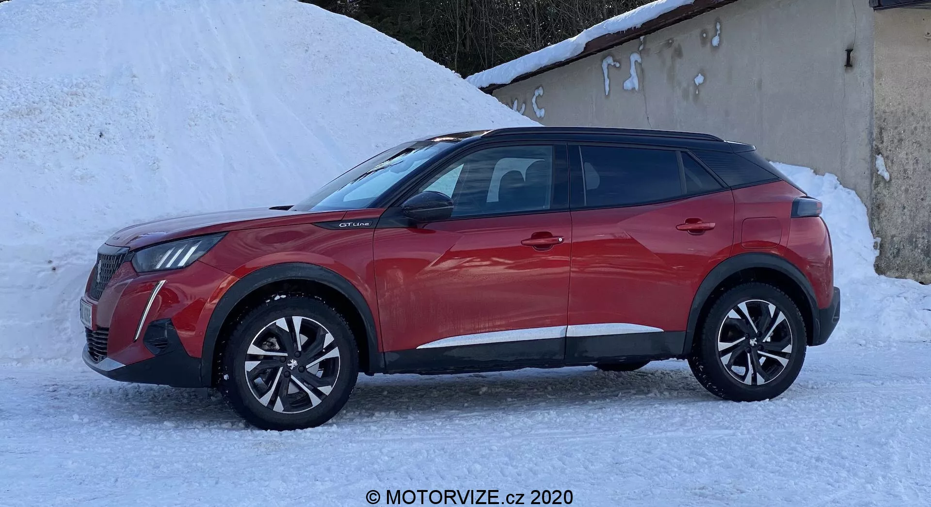 Widok boczny nowoczesnego SUV-a Peugeot 2008 z 2020 roku, w jasnej i błyszczącej czerwonej farbie. Pojazd jest ustawiony na zaśnieżonym podłożu, co sugeruje przydatność do zimowych warunków. Dynamiczny i kanciasty design charakteryzuje się wyraźnymi łukami kół, ostrymi liniami nadwozia, opadającą linią dachu, felgami aluminiowymi i lusterkami bocznymi w kolorze nadwozia z możliwością zaawansowanych funkcji, takich jak regulacja elektryczna lub monitorowanie martwego pola. Czterodrzwiowy układ z przyciemnianymi szybami, czarne dolne listwy ochronne i skomplikowane felgi podkreślają zarówno sportową, jak i wytrzymałą estetykę.