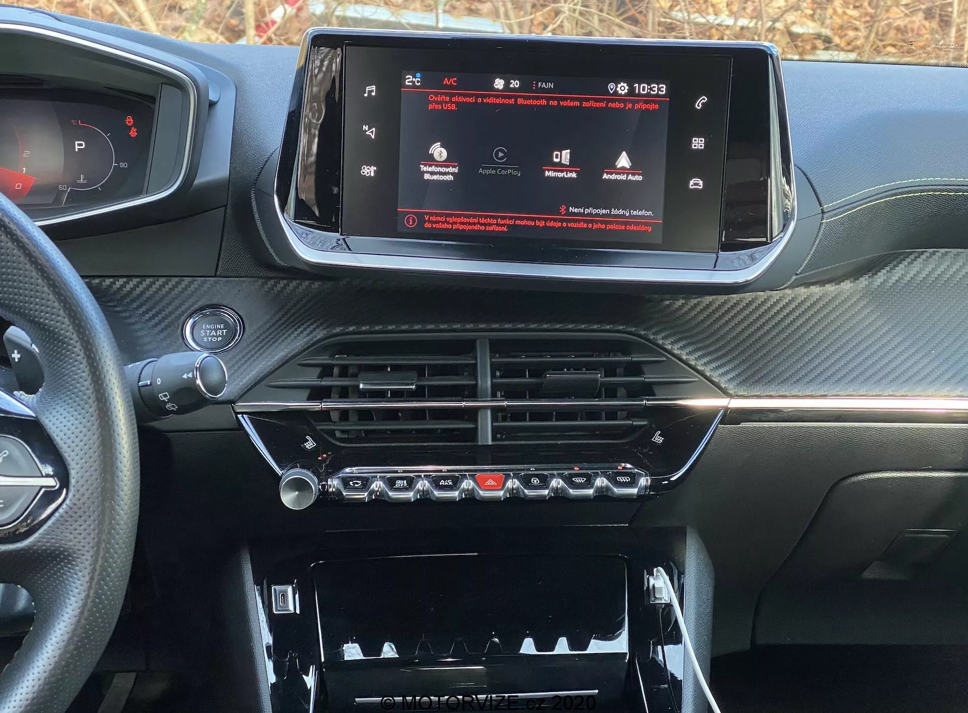 Zbliżenie wnętrza deski rozdzielczej nowoczesnego samochodu, konkretnie Peugeot 2008 2020 (peugeot-2008-1-2-puretech-100-active). Zdjęcie zrobione z perspektywy kierowcy lub przedniego pasażera, skoncentrowane na konsoli centralnej i systemie infotainment. Panoramiczny ekran dotykowy pokazuje menu ustawień pojazdu, wskazując zaawansowaną technologię. Poniżej znajdują się chromowane nawiewy i sterowanie klimatyzacją z fizycznymi przyciskami i pokrętłami. Niżej widoczny jest schowek z pokrywą. Deska rozdzielcza ma teksturowane wykończenie podobne do włókna węglowego, nadając sportowy i luksusowy wygląd.