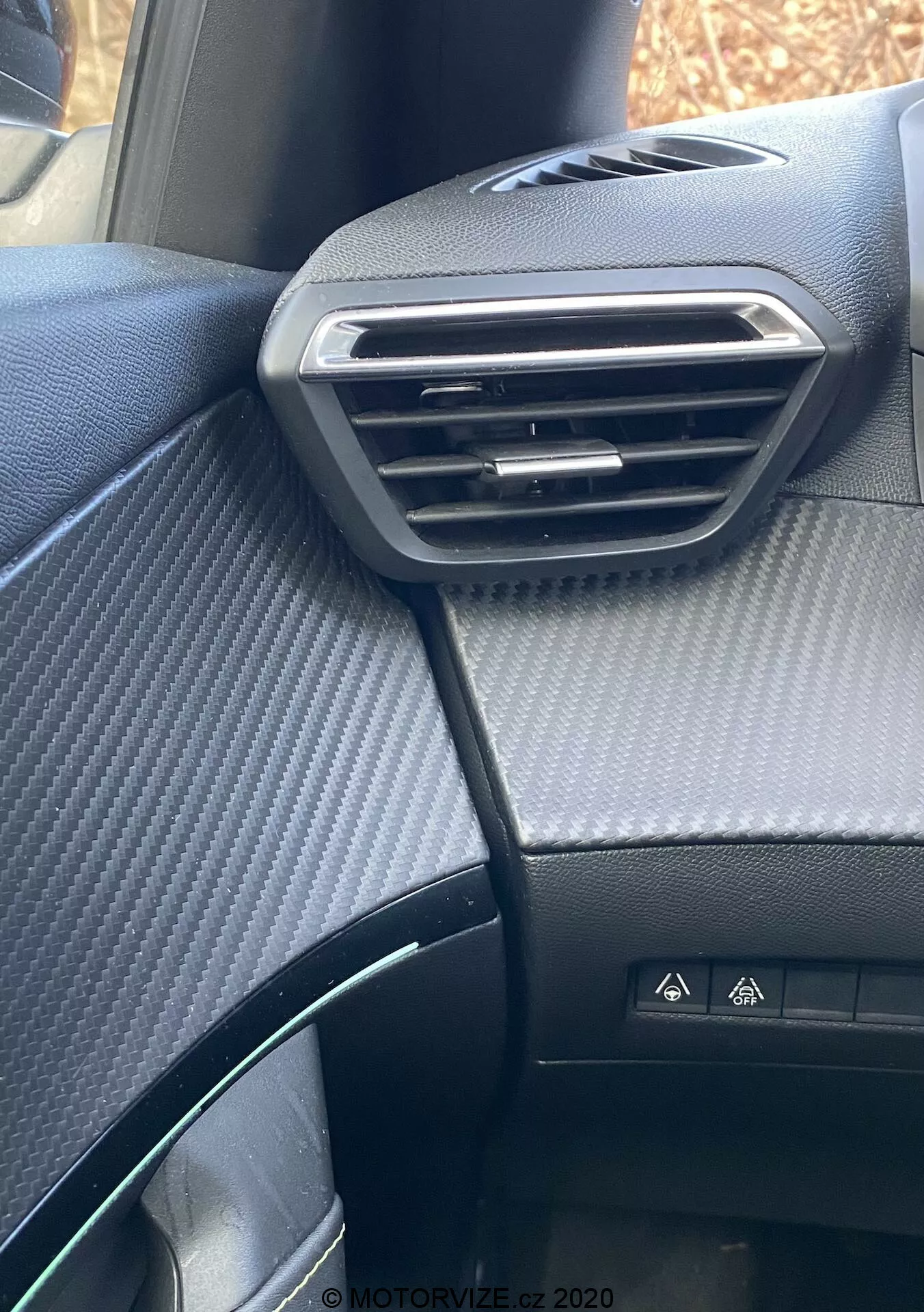 Zbliżenie na deskę rozdzielczą po stronie pasażera we wnętrzu Peugeot 2008 2020, podkreślające srebrny, regulowany nawiew, teksturowaną górną część deski rozdzielczej imitującą włókno węglowe, przycisk świateł awaryjnych, częściowo widoczny kierownicę oraz czarne siedzenie w kropki.