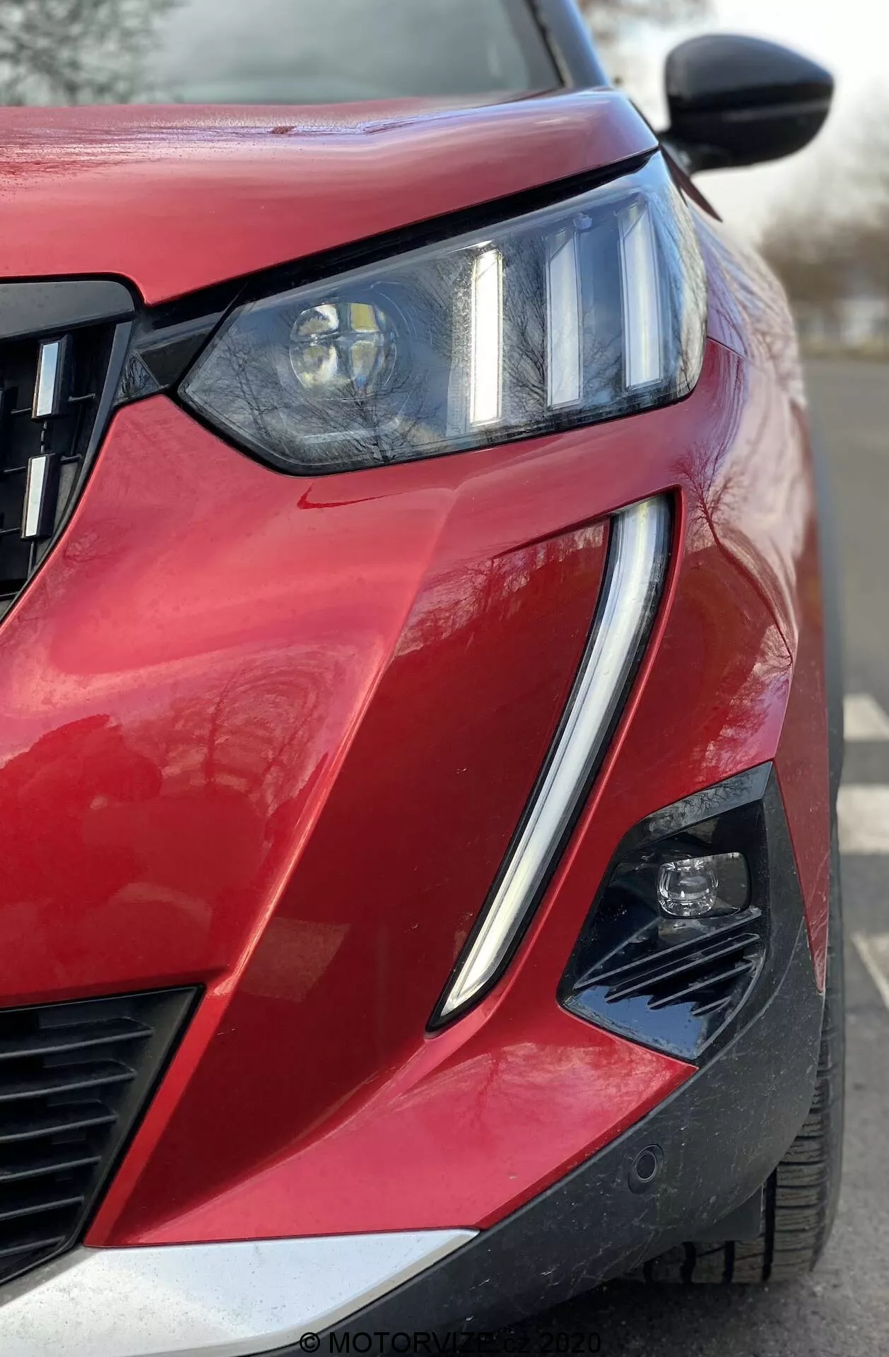 Zbliżenie na frontalny widok pod kątem czerwonego Peugeot 2008 1.2 PureTech 100 Active z 2020 roku, ukazujący przedni lewy kwartał. Wyraźny reflektor z belką projektora i zintegrowanymi LED DRL, pozioma krata wlotu powietrza oraz częściowo widoczny zespół drugiego światła w kanciaście wyciętym zderzaku. Błyszcząca farba z metalicznym wykończeniem, ostra linia nadwozia, podniesiony kontur maski, czarne lusterko boczne prawdopodobnie ze zintegrowanymi kierunkowskazami lub kamerą martwego pola oraz wycięcie w dolnym rogu zderzaka sugerujące zaawansowane systemy wspomagania kierowcy.